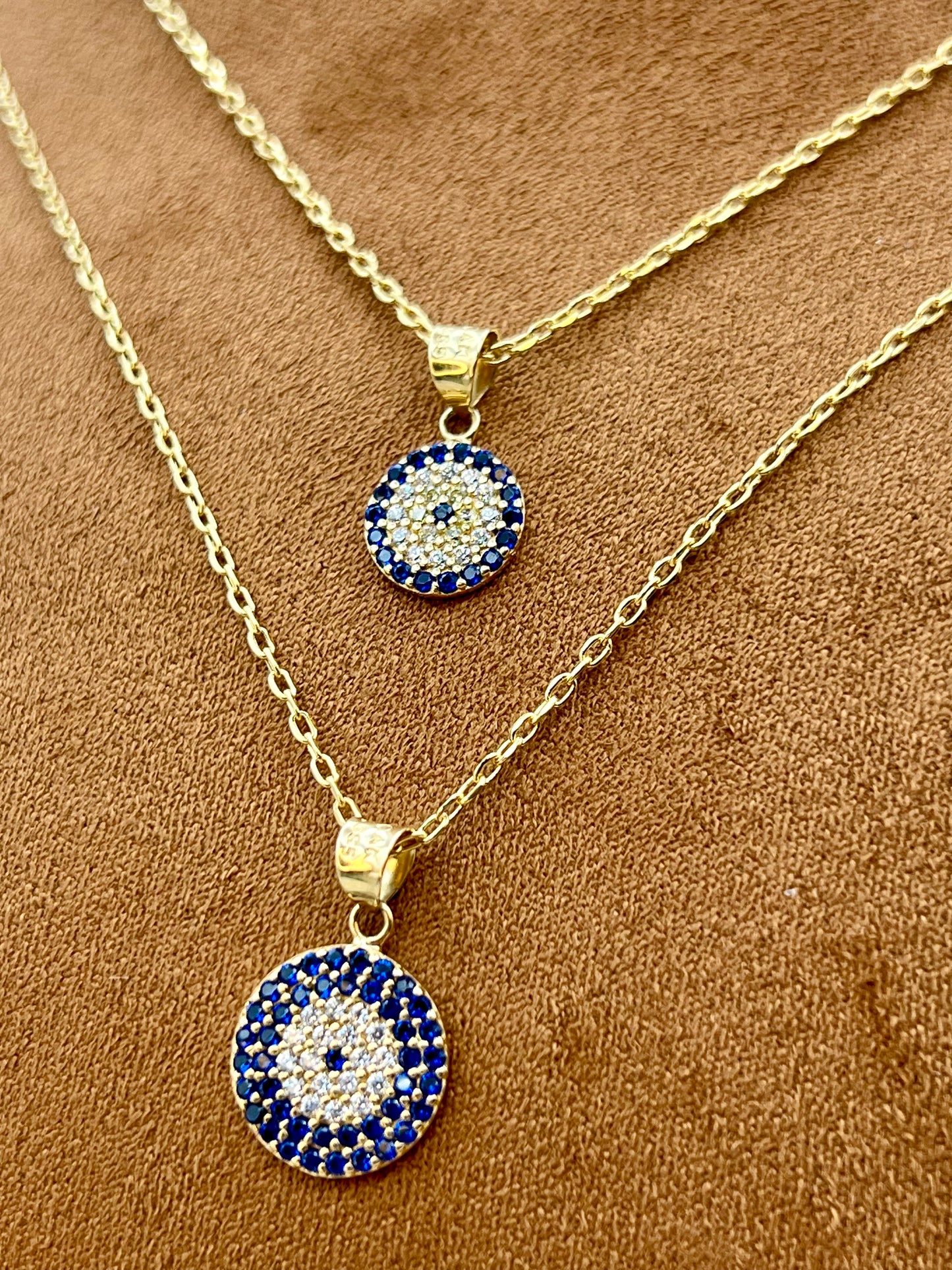 Two gold chain necklaces with circular blue and white evil eye pendants, displayed on a brown textured fabric background.
