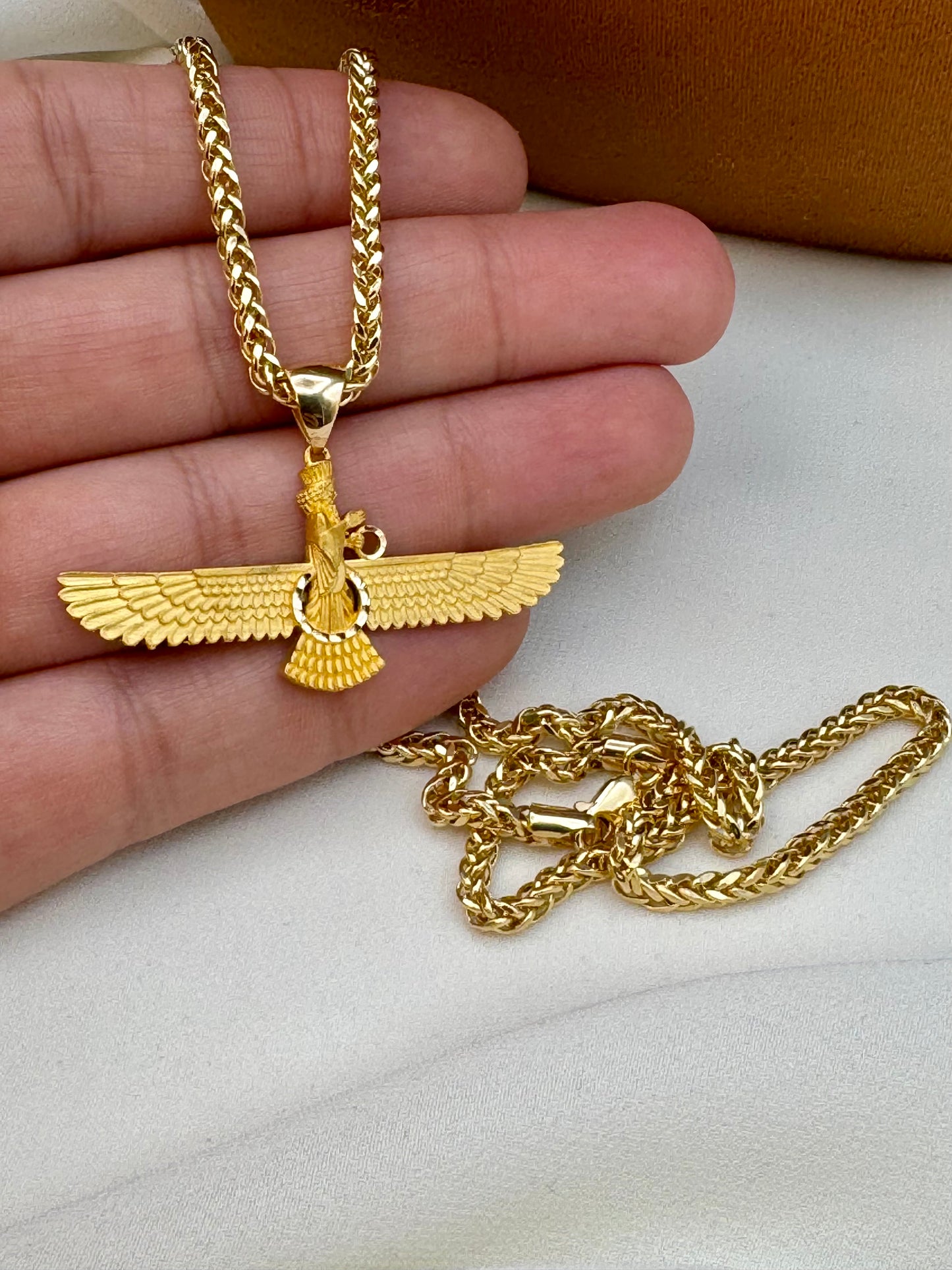 farvahar Gold necklace with an eagle pendant held by a hand on a white surface.