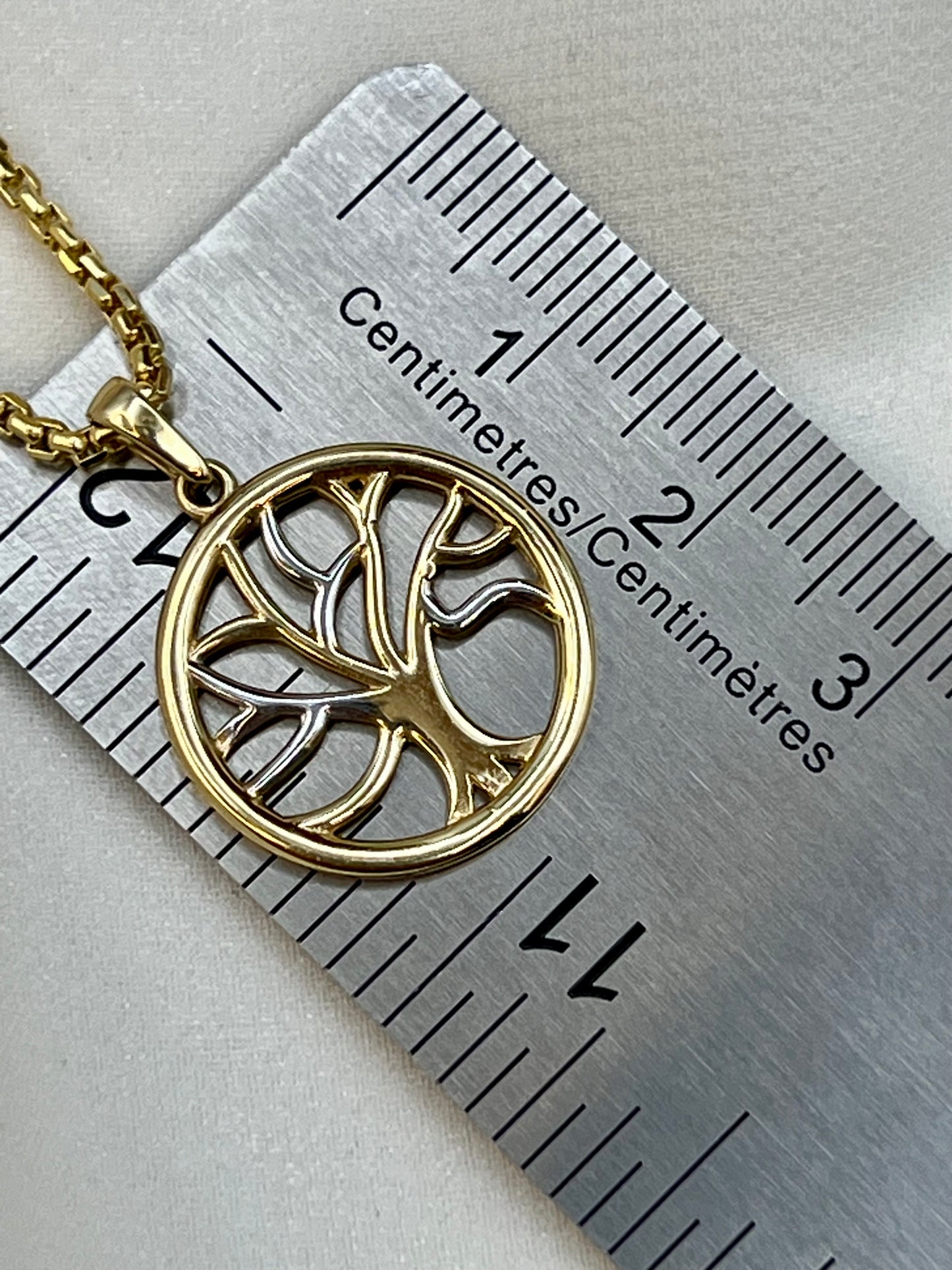 A gold pendant necklace with a tree of life design inside a circle, shown next to a metal ruler for scale on a light gray surface.