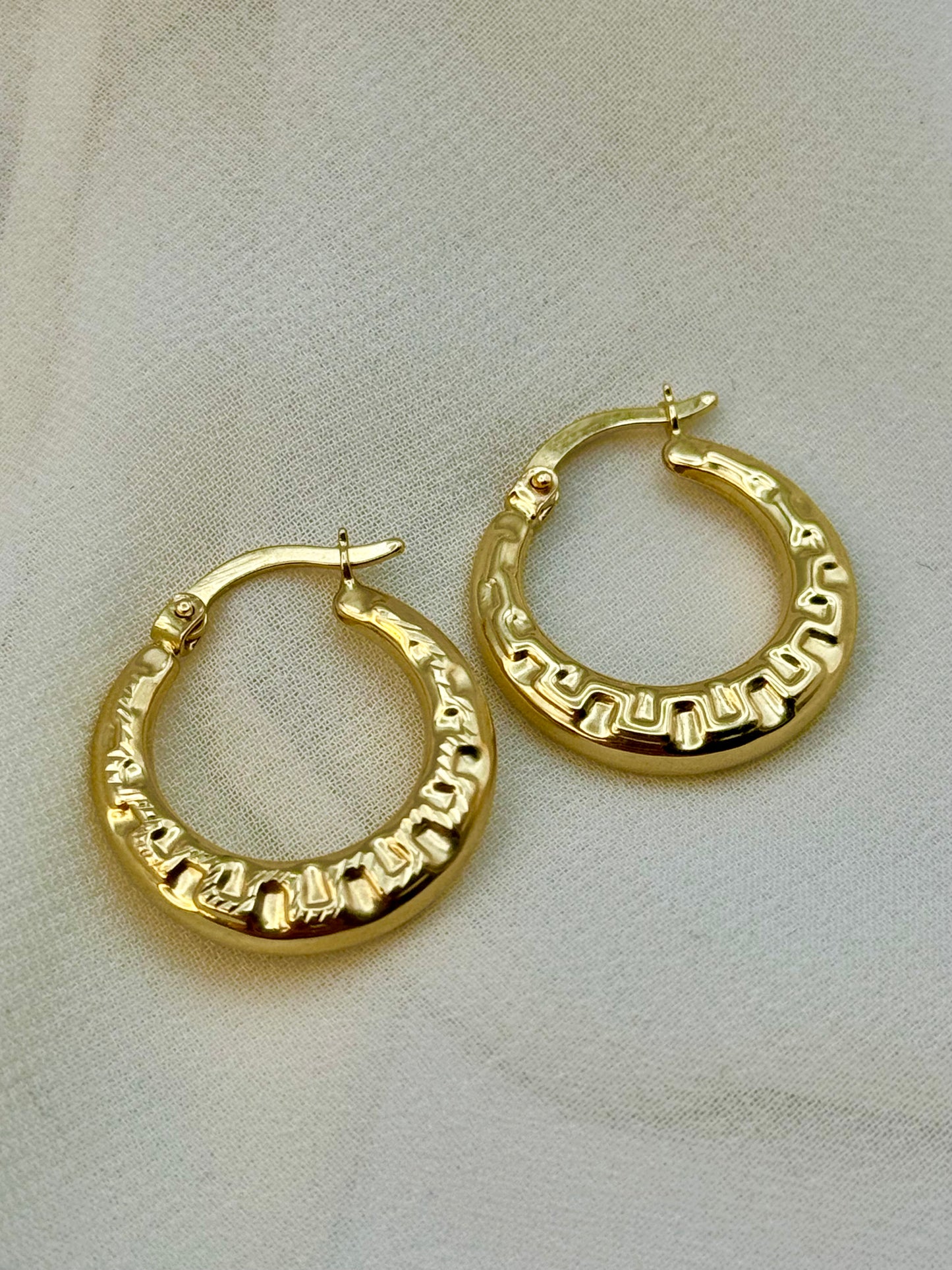 A pair of 14k gold fat hoop earrings with a hollow design, displayed on a white fabric surface.