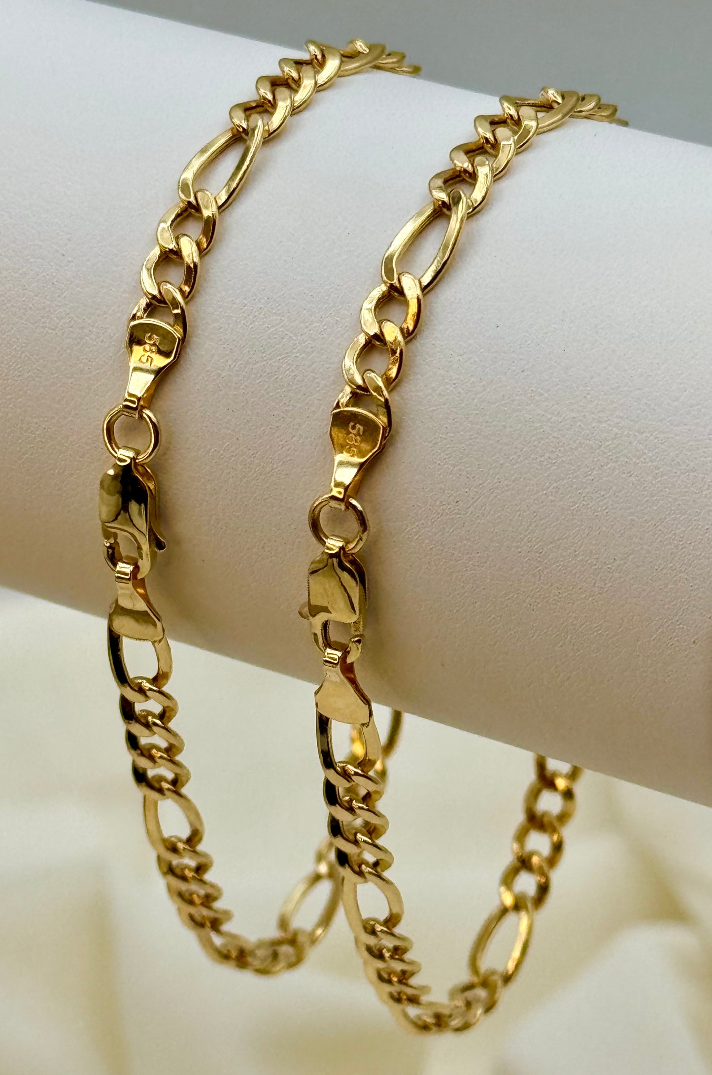 Two 14k gold figaro chain bracelets on a white arm against a beige background