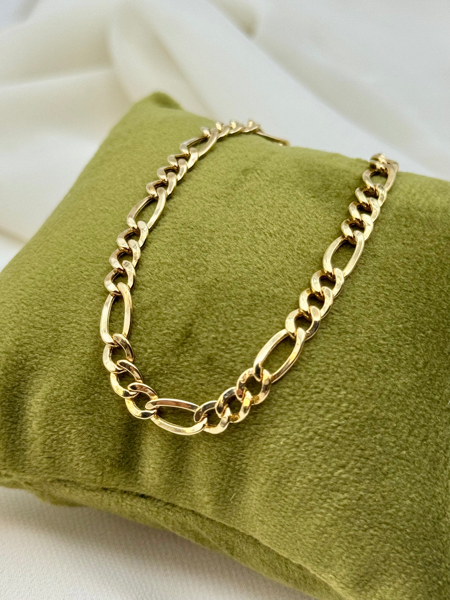 Gold chain figaro bracelet on a green cushion with a white background