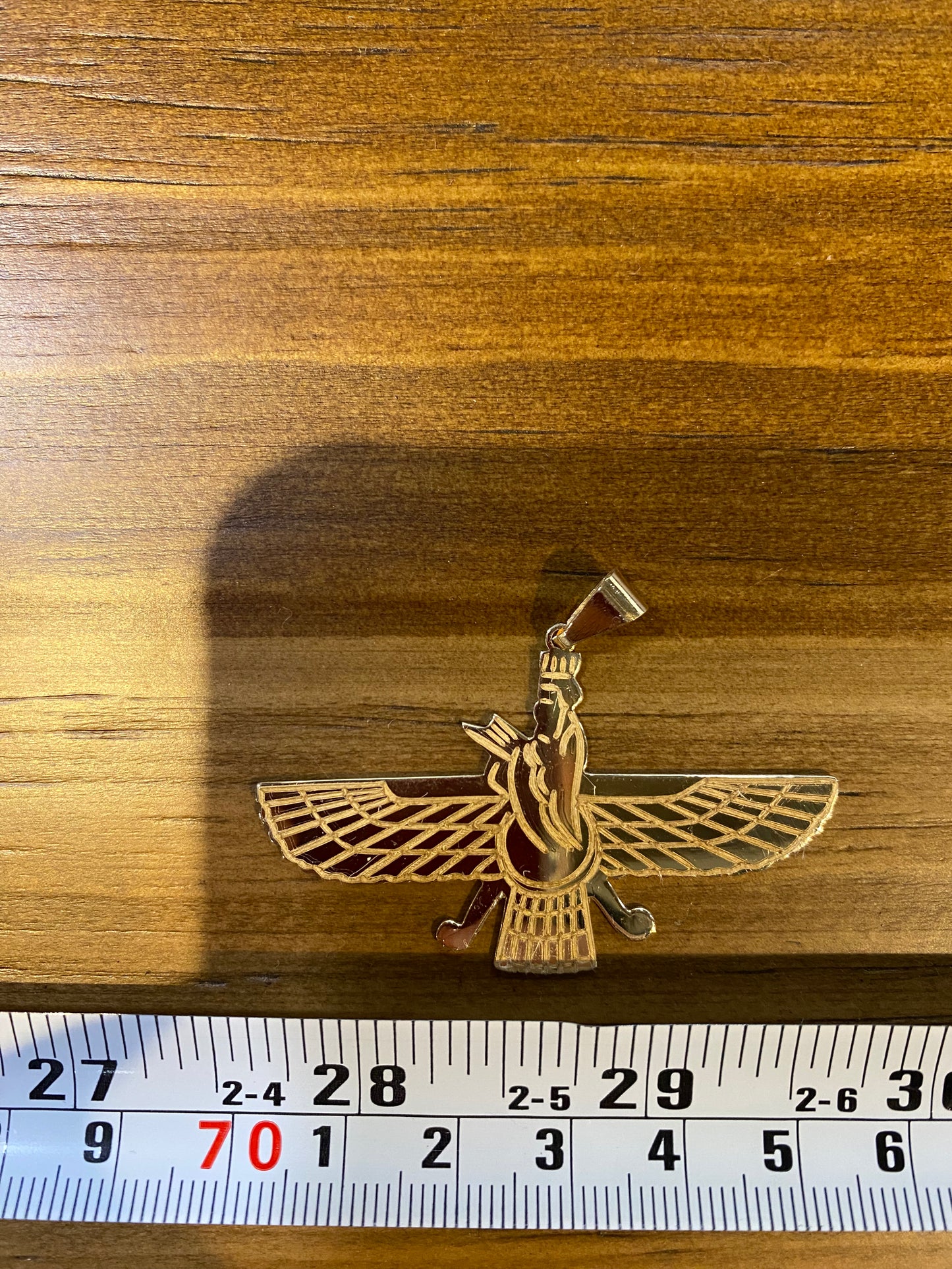Gold-colored Faravahar symbol pendant on a wooden surface with a ruler for scale.