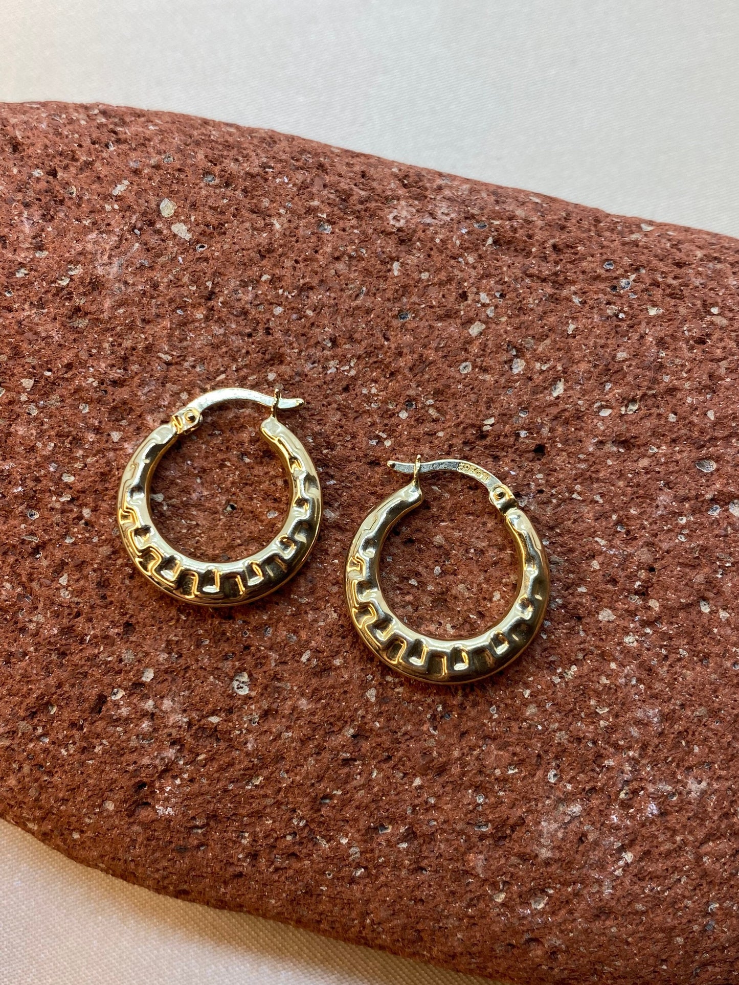 A pair of 14k gold hoop earrings with a Greek design, displayed on a textured surface.