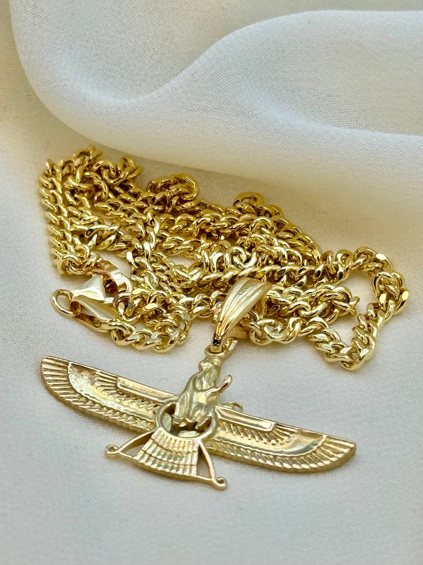 An 18K gold necklace featuring the Faravahar symbol, which is a Zoroastrian symbol representing Ahura Mazda, the supreme god. The necklace is displayed on a white fabric surface.