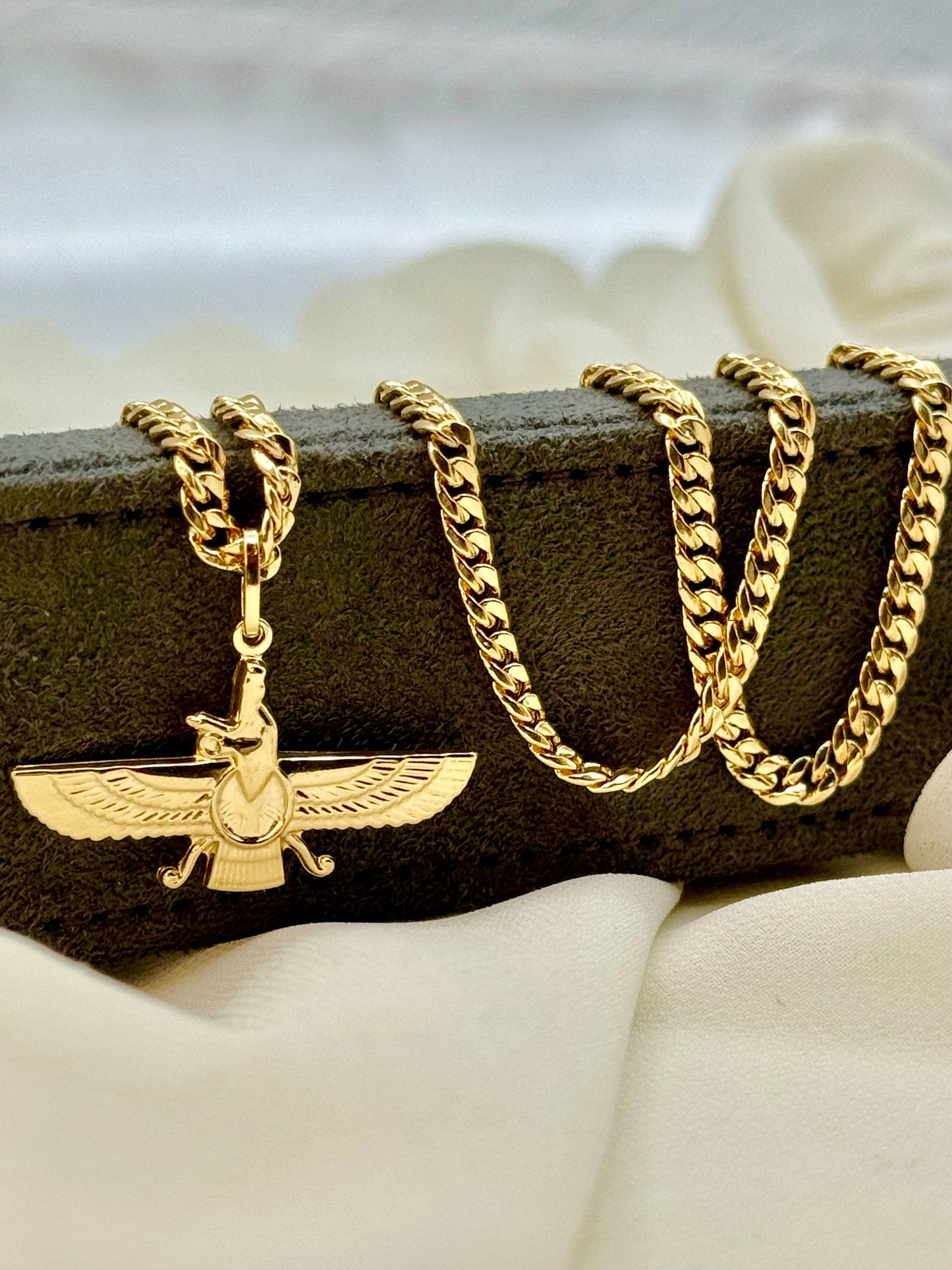 An 18k gold Faravahar necklace displayed on a white fabric surface, featuring a curb chain design.