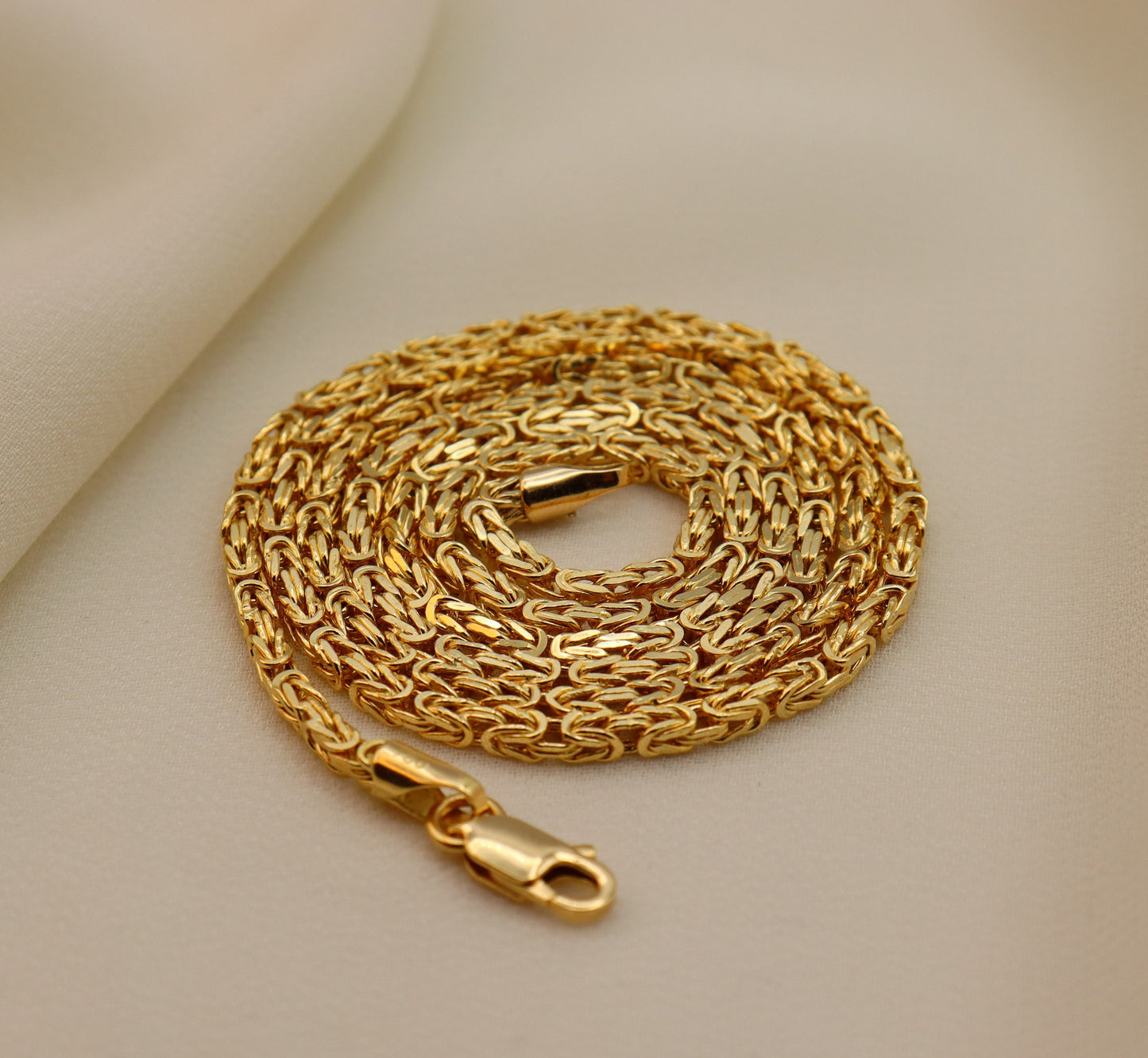 A 18K gold Byzantine chain necklace with a secure lobster claw closure, displayed on a beige background.