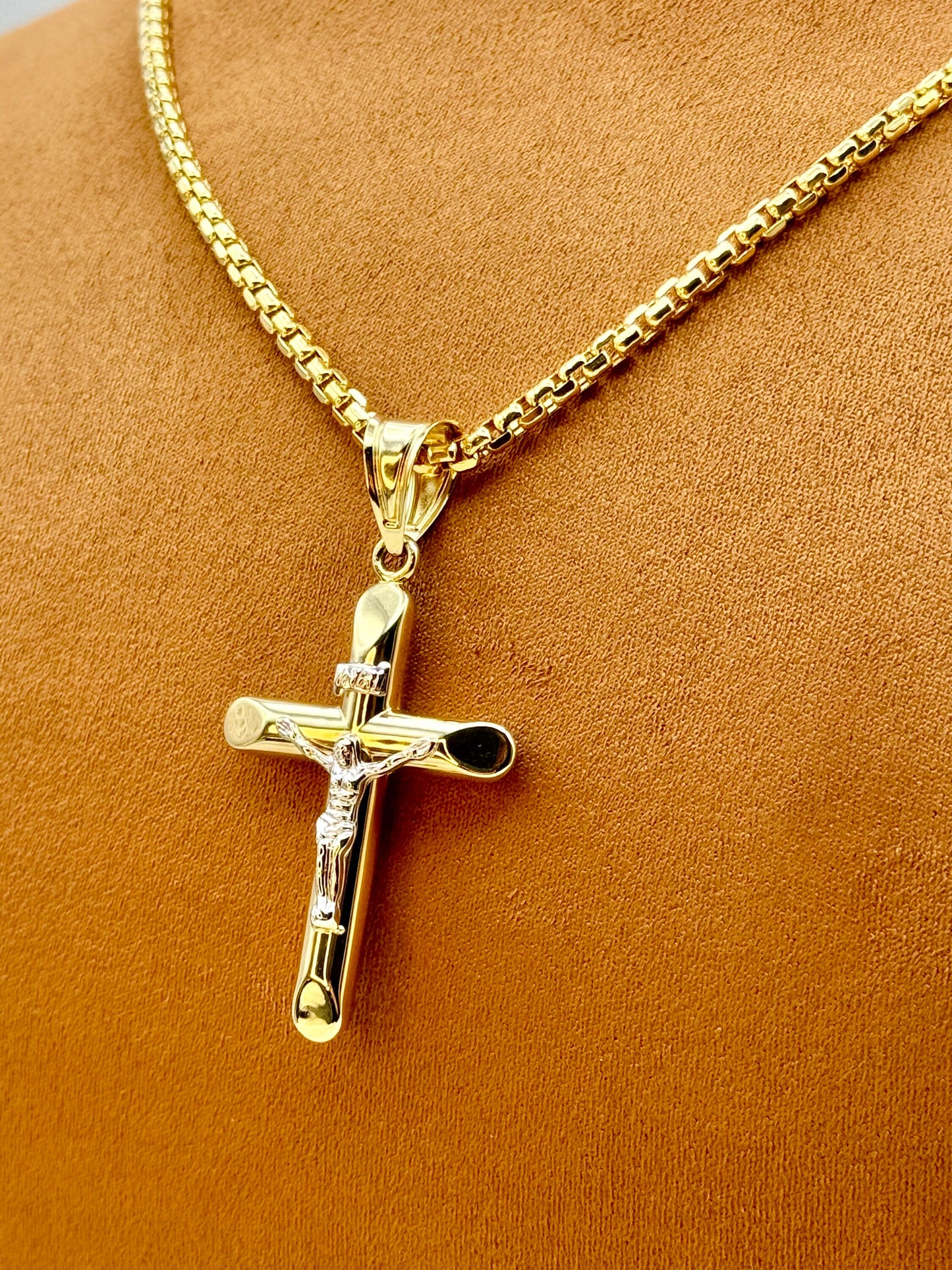 The image displays a 14K gold cross necklace with a crucifix pendant, featuring a box chain design. The necklace is presented on a mannequin with a warm brown background.