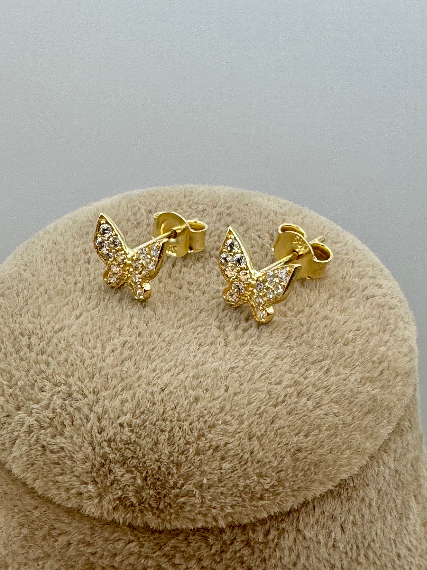 A pair of gold butterfly stud earrings with clear pavé-set stones, displayed on a round beige textured jewelry stand against a neutral gray background.