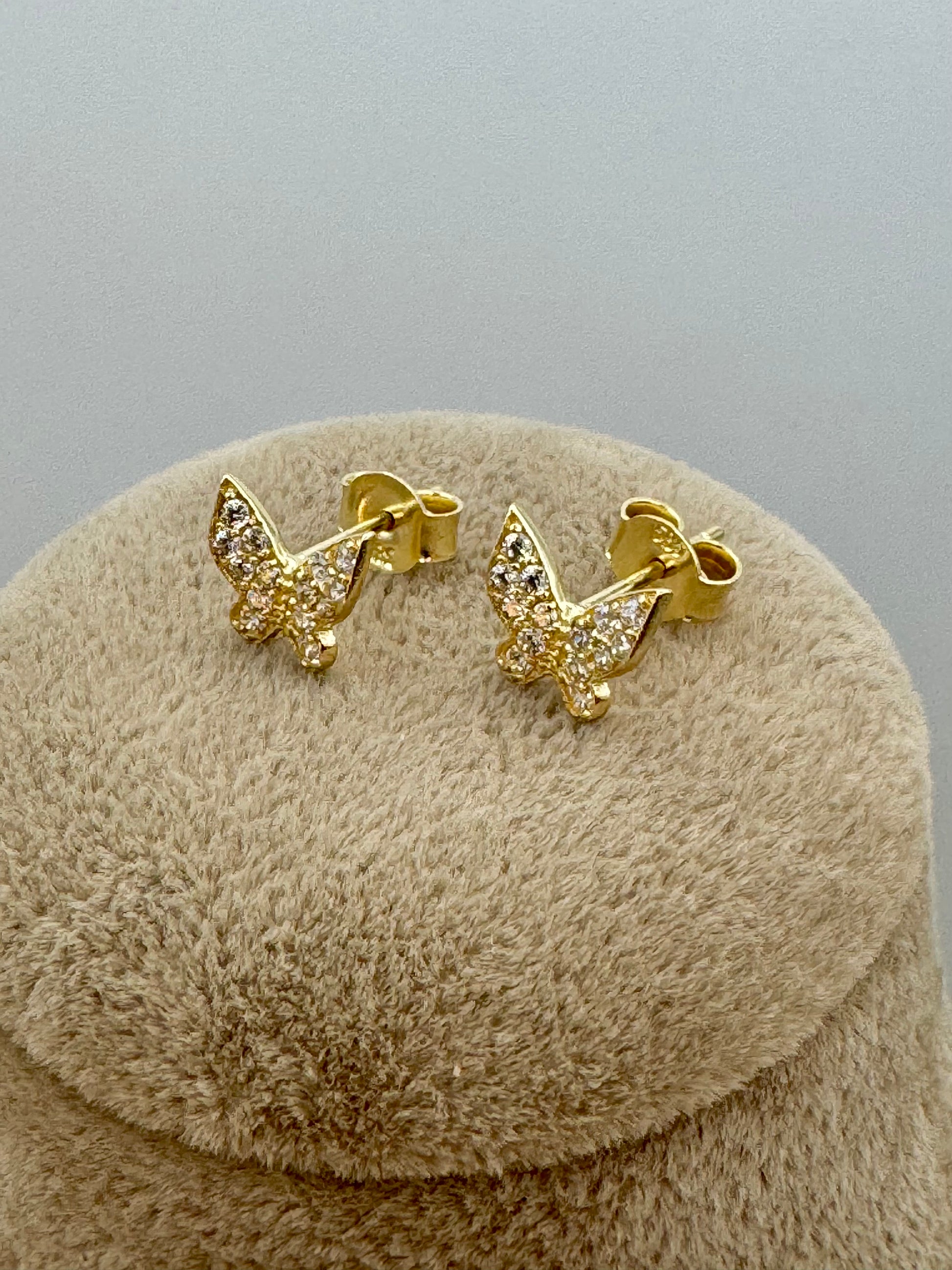 A pair of gold butterfly stud earrings with clear pavé-set stones, displayed on a round beige textured jewelry stand against a neutral gray background.