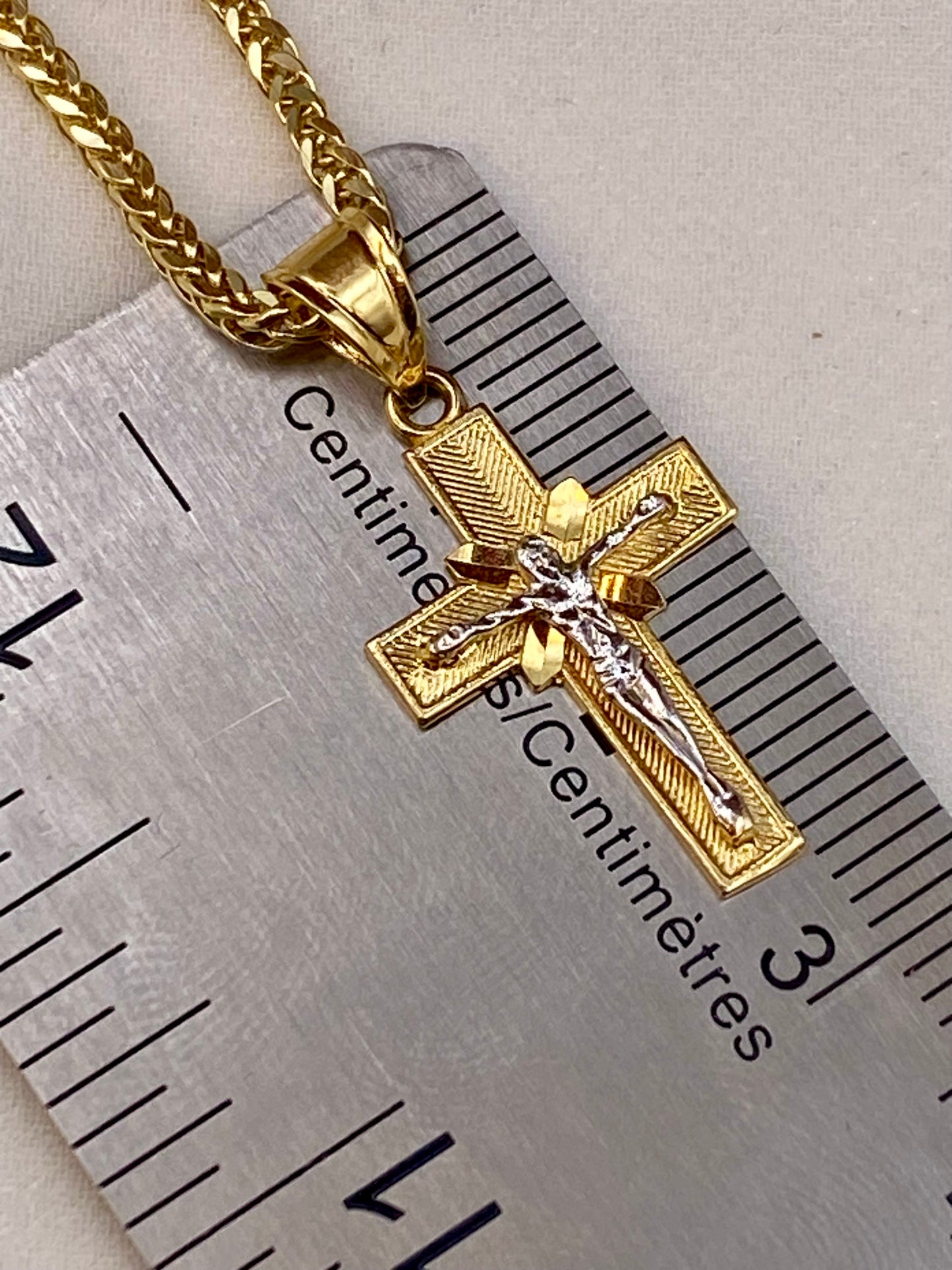 Gold cross pendant necklace with a silver Jesus figure, shown next to a metal ruler for scale on a neutral background.