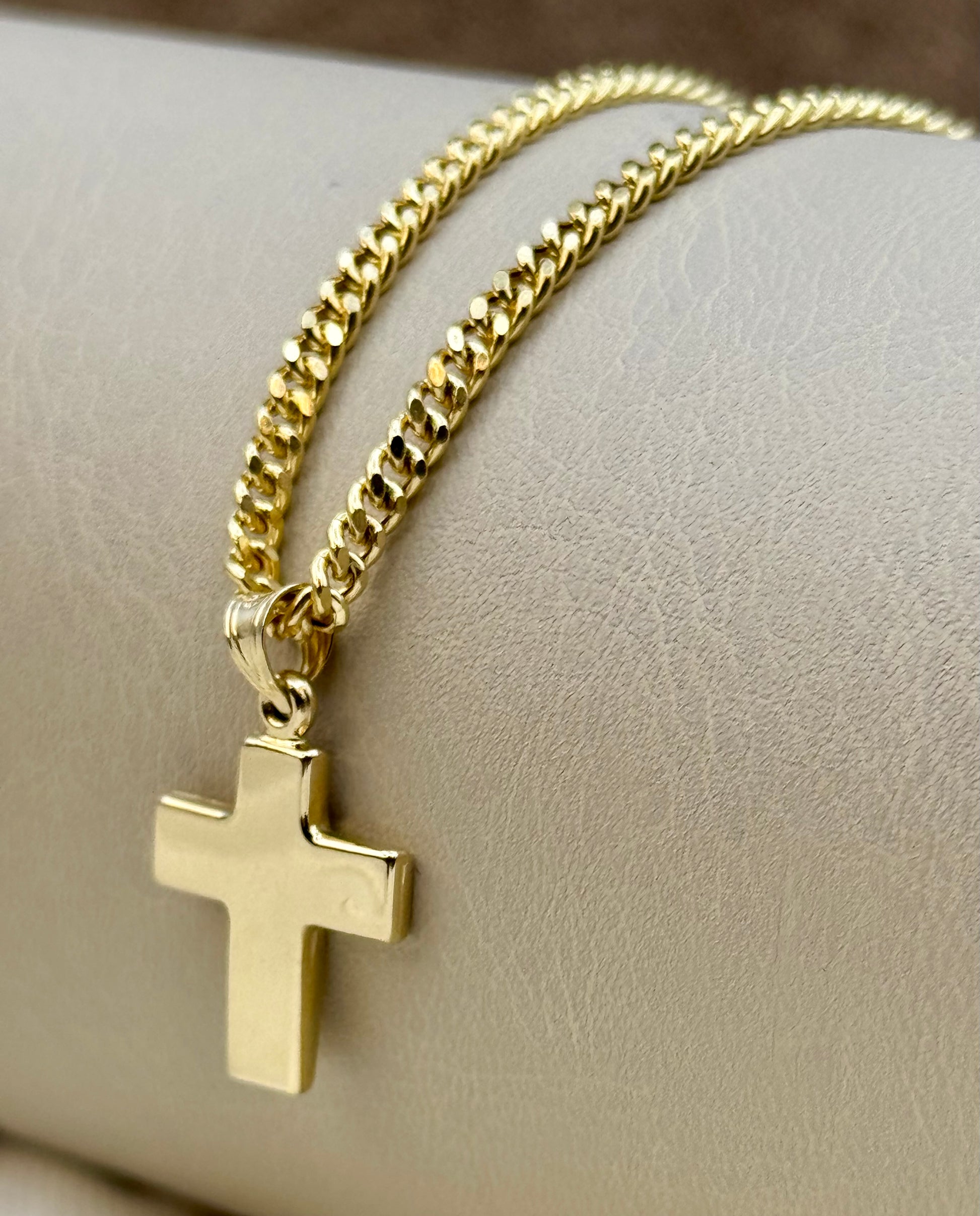 A gold-toned cross pendant necklace on a curb chain, displayed on a textured beige jewelry stand.