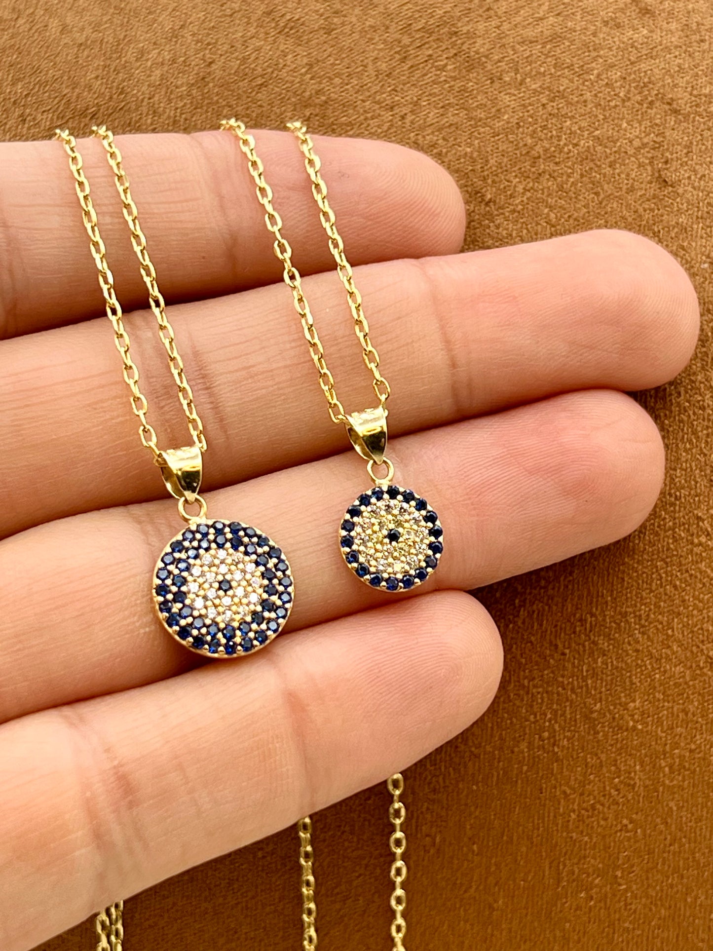 Gold-tone circular pendant necklace and matching earrings with a dark blue and gold evil eye design, displayed on a person's hand against a brown textured background.