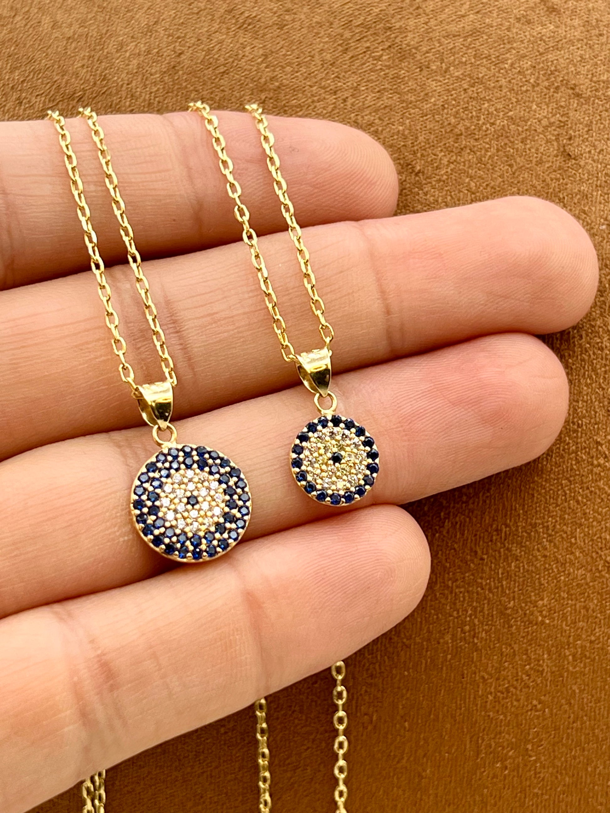 Gold-tone circular pendant necklace and matching earrings with a dark blue and gold evil eye design, displayed on a person's hand against a brown textured background.