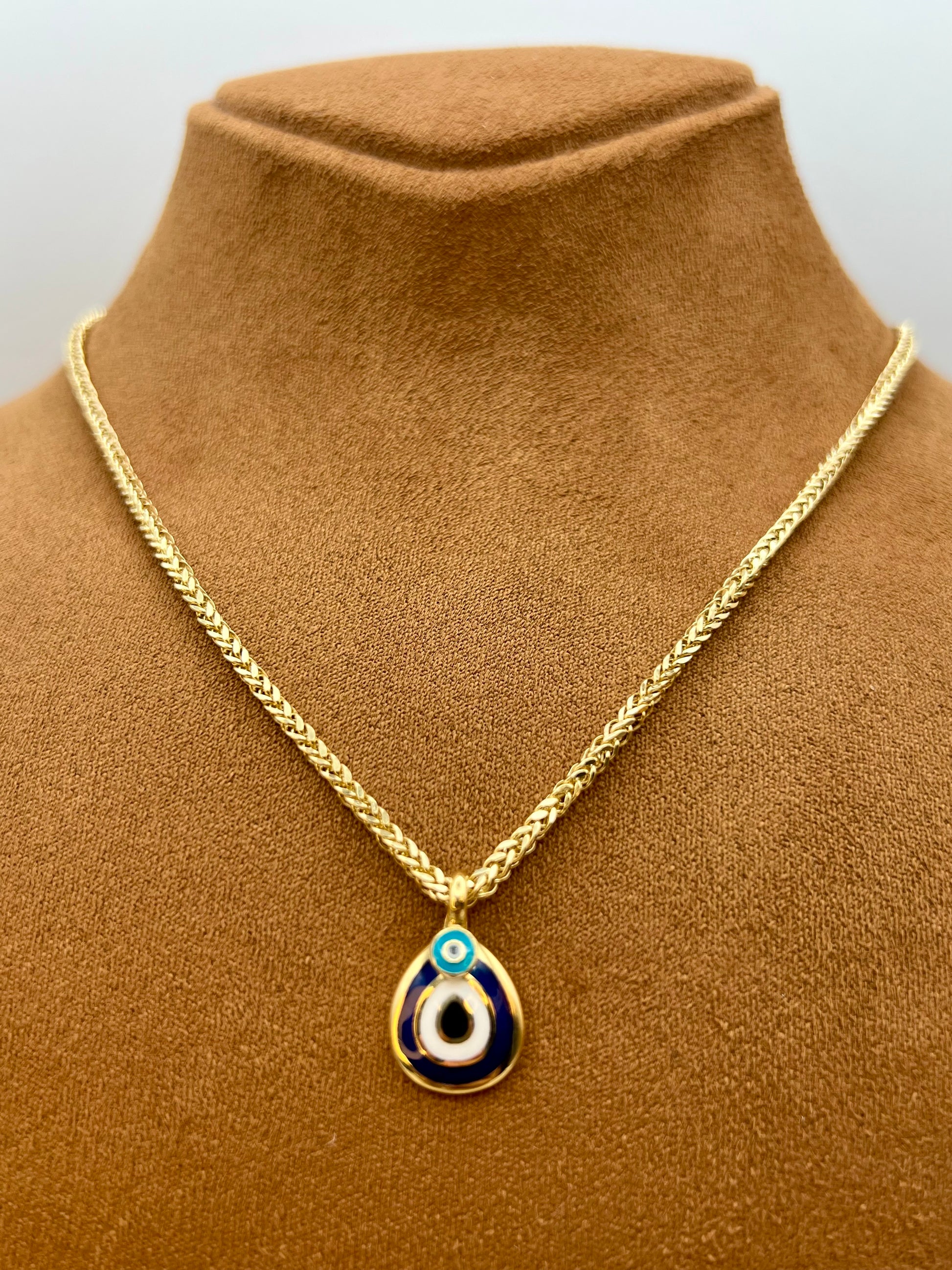A gold chain necklace with a teardrop-shaped evil eye pendant, displayed on a brown velvet jewelry bust. The pendant features blue, white, and light blue enamel details.