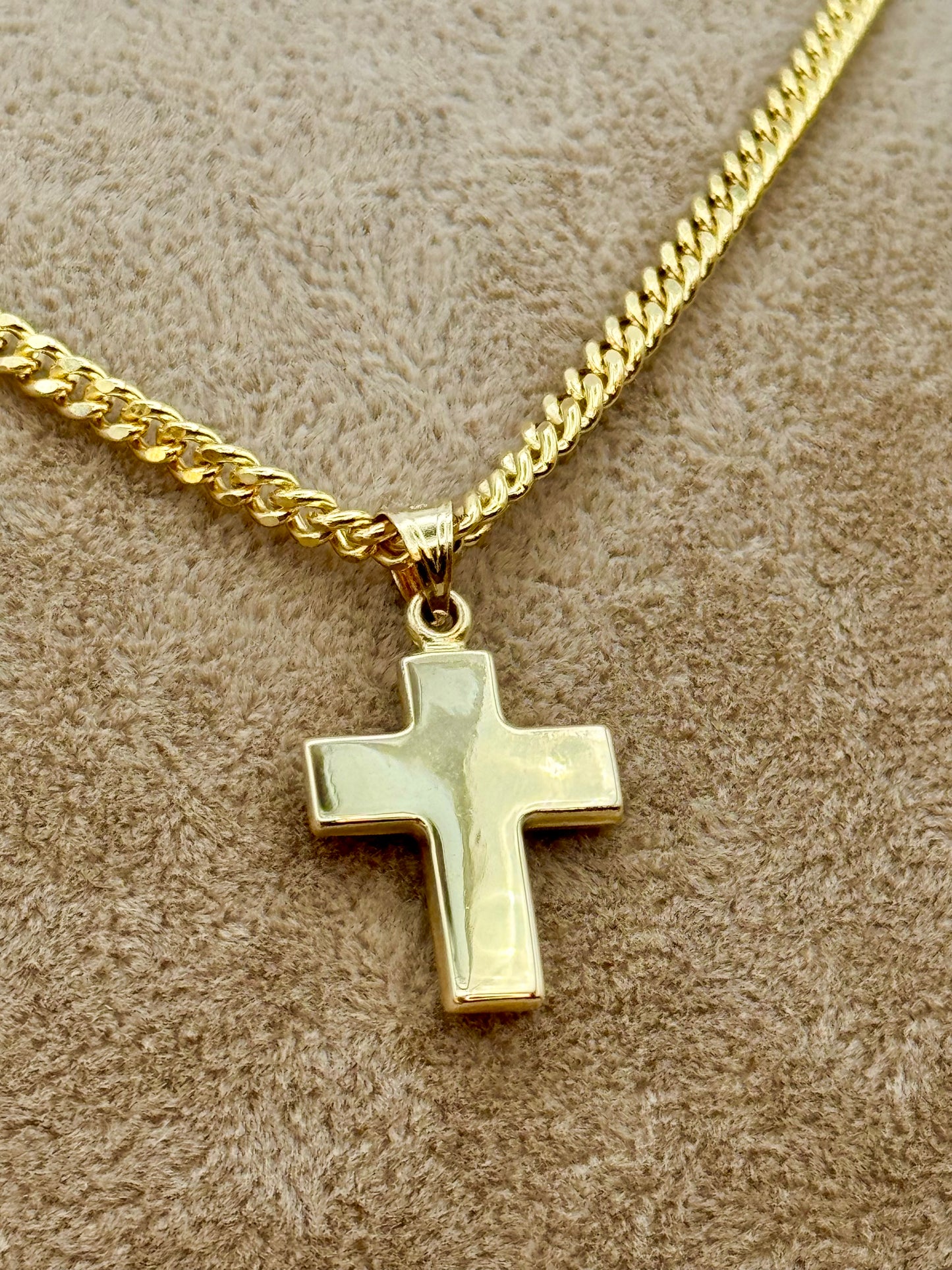 A gold cross pendant necklace on a gold chain, displayed on a textured beige surface.
