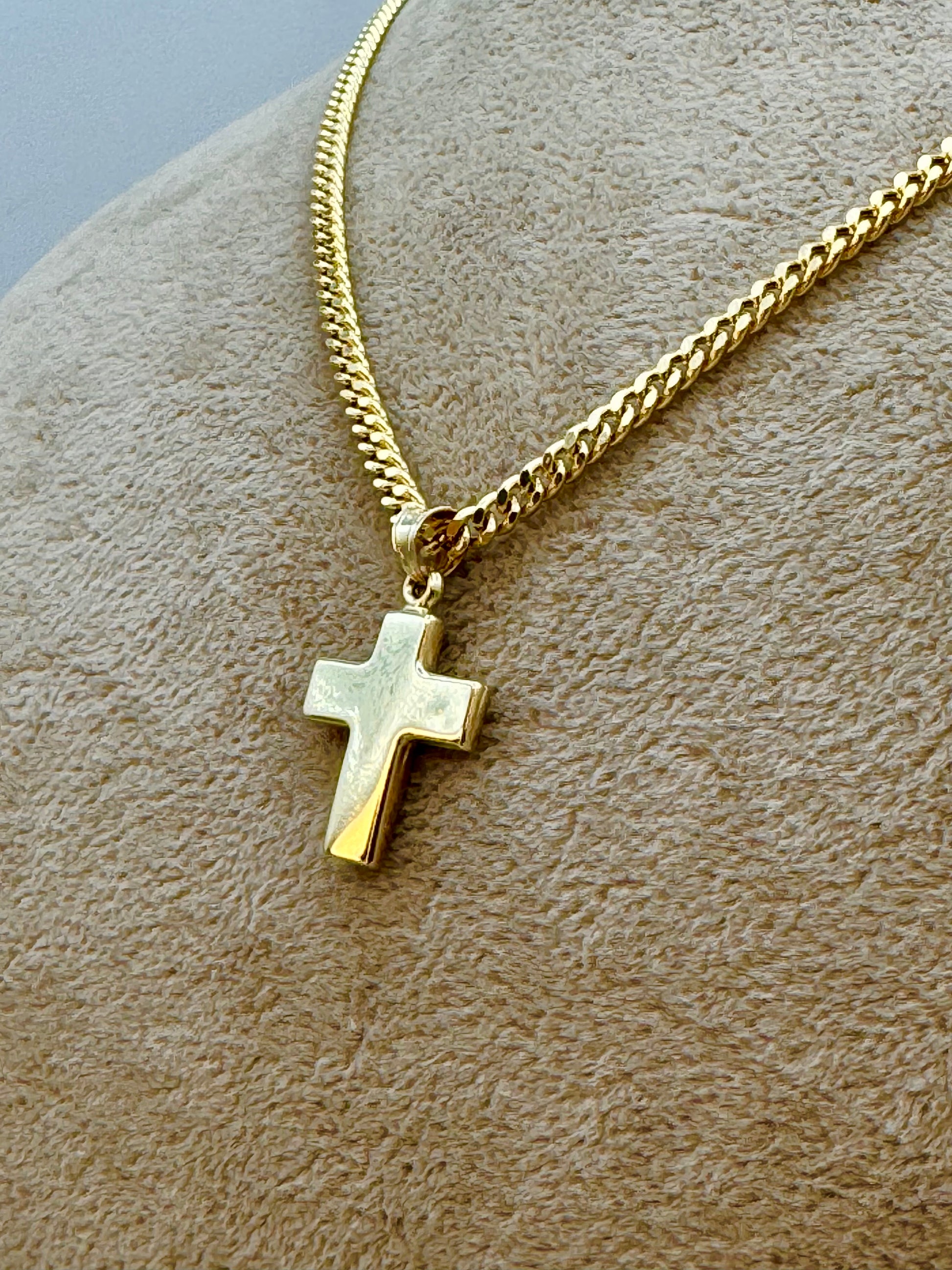 A gold cross pendant necklace on a gold curb chain, displayed on a textured beige jewelry bust.