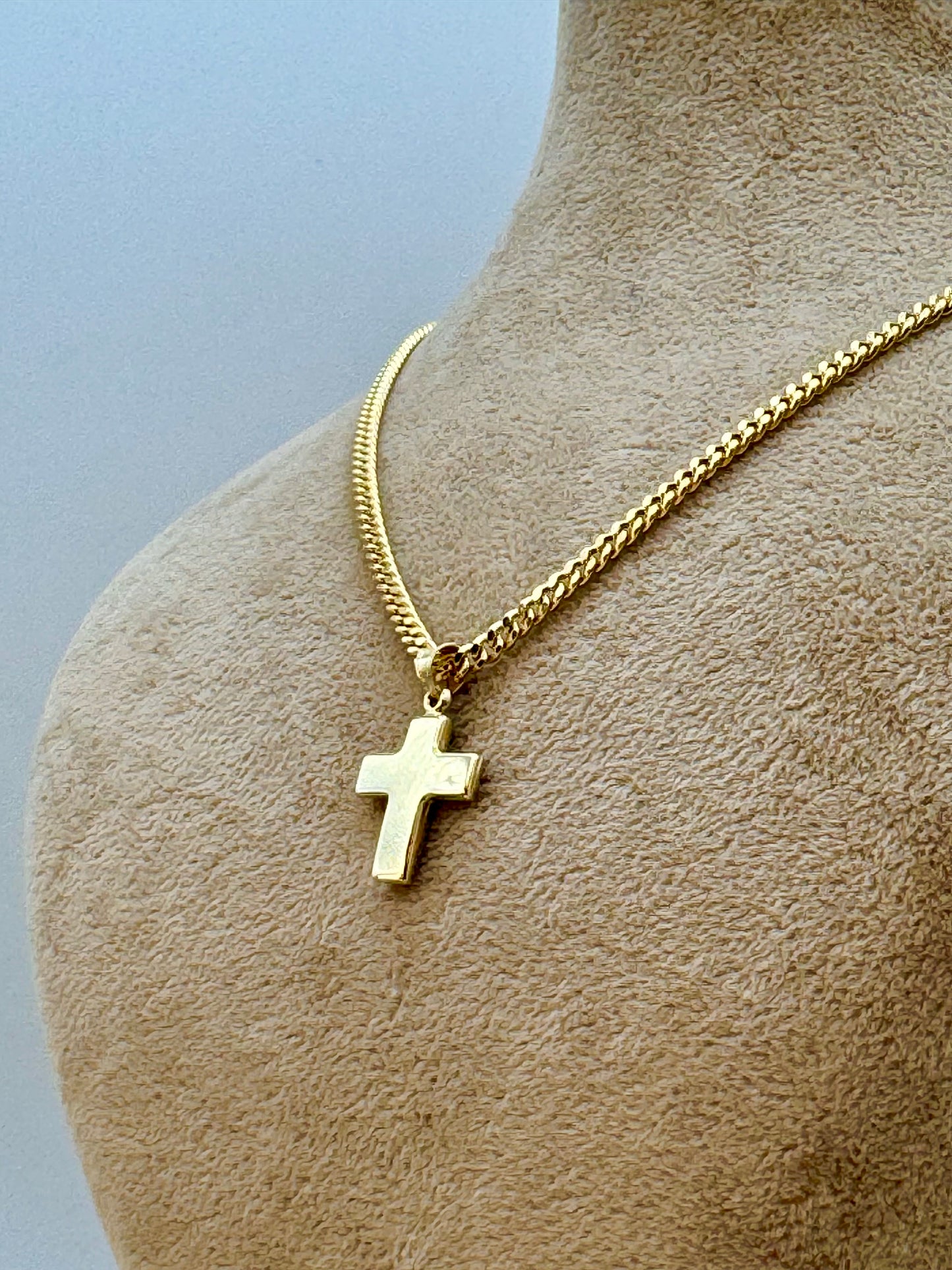 A gold cross pendant necklace displayed on a beige fabric mannequin bust against a light blue-gray background.