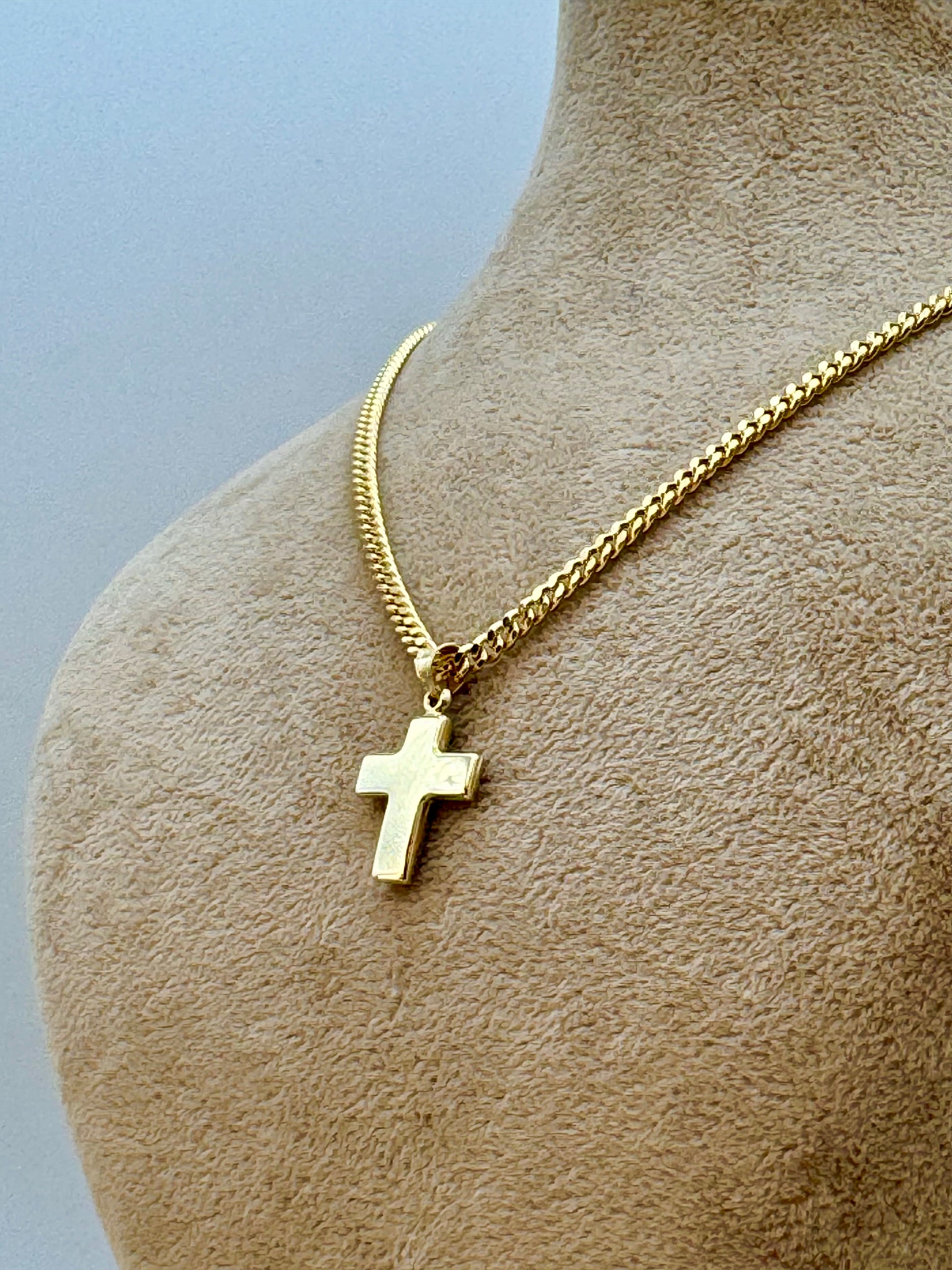 A gold cross pendant necklace displayed on a beige fabric mannequin bust against a light blue-gray background.