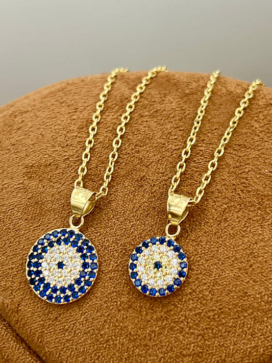 Two gold necklaces with circular evil eye pendants displayed on a brown textured surface. Each pendant features a white and blue concentric eye design with blue rhinestones.