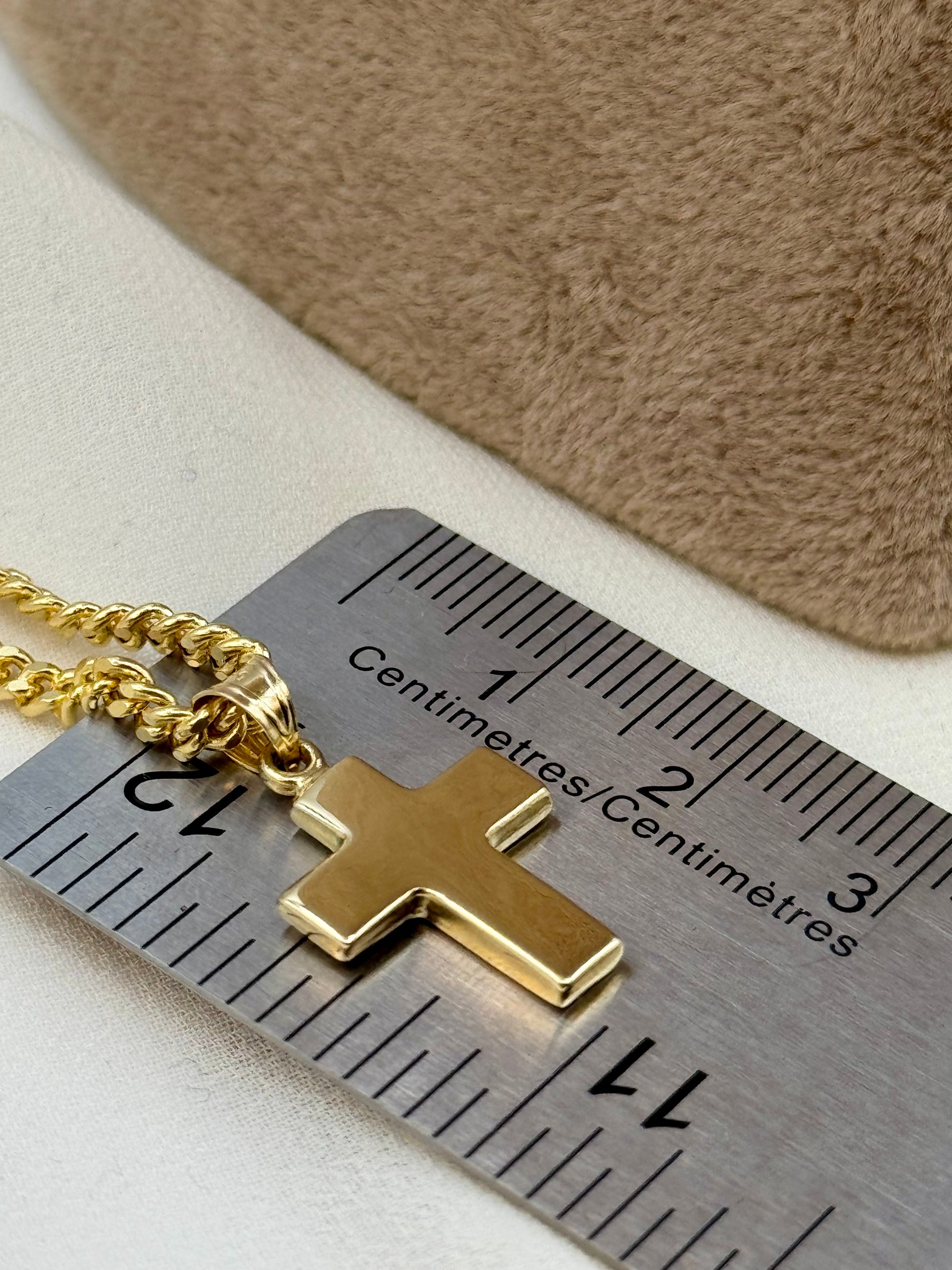 A gold cross pendant necklace on a gold chain, shown next to a metal ruler for scale with a beige textured background.