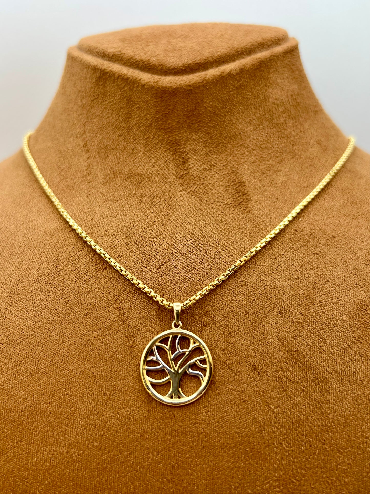 A gold tree of life pendant necklace displayed on a brown velvet jewelry bust against a light gray background.