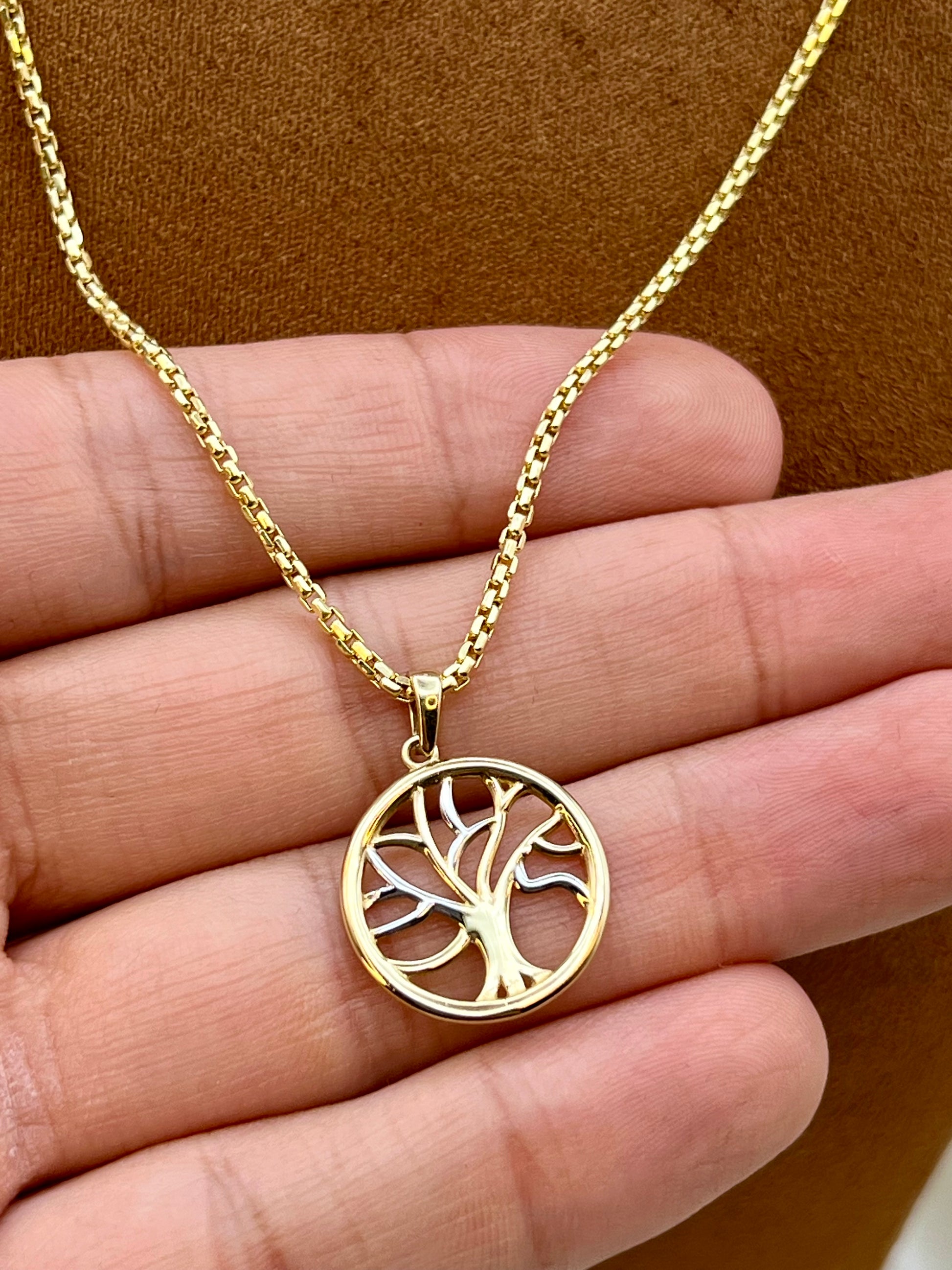A gold tree of life pendant necklace held in a hand against a brown textured background.