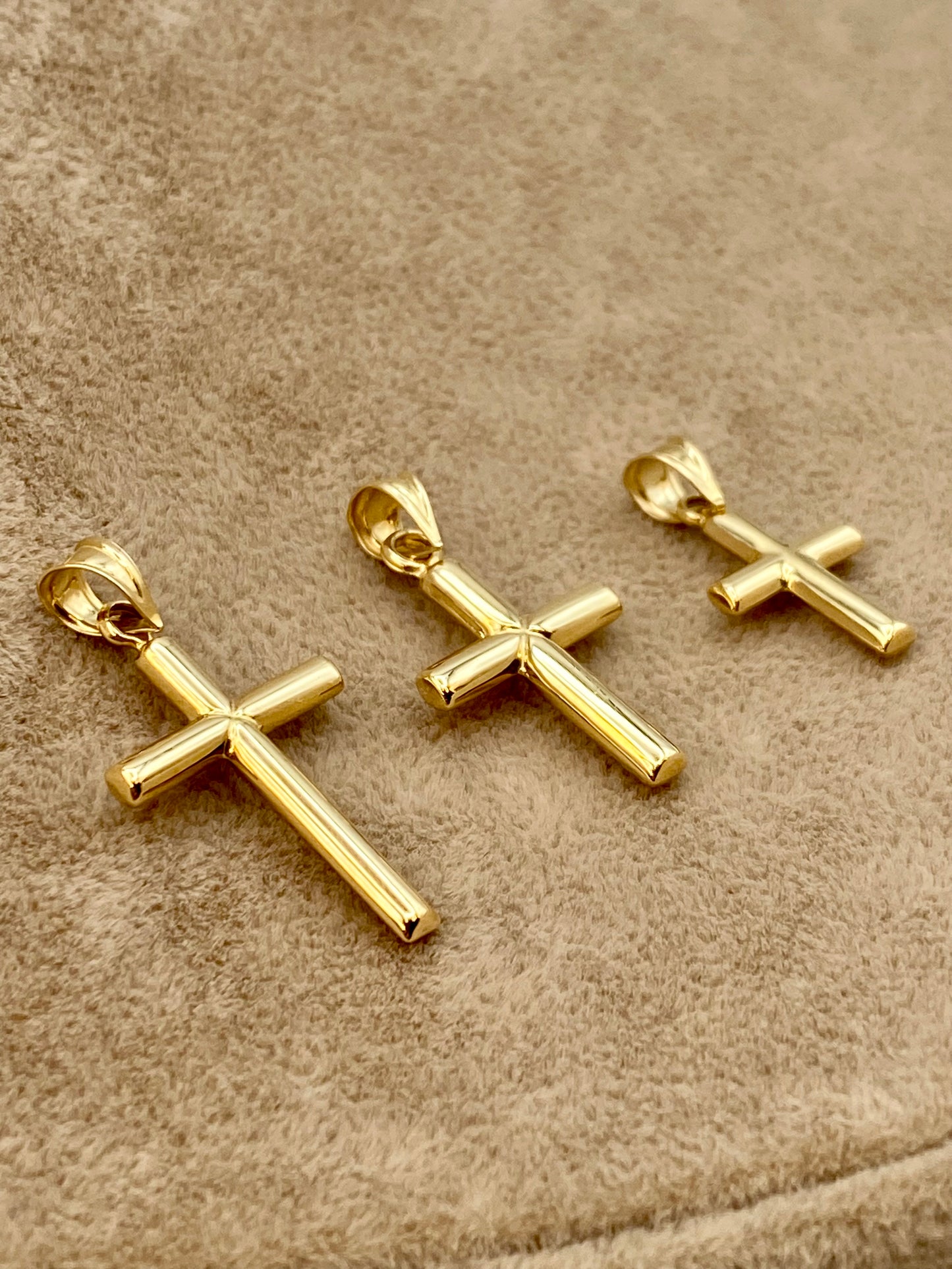 Three gold cross pendants with polished metallic finishes, arranged diagonally on a textured beige fabric background.