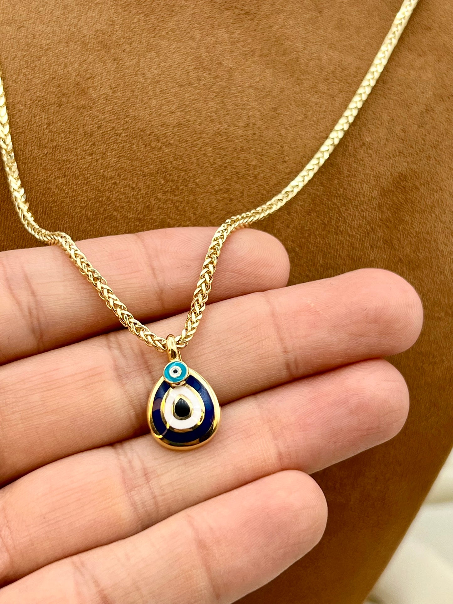 A gold chain necklace with a teardrop-shaped evil eye pendant, held in a hand against a brown textured background. The pendant features a gold border, a dark blue enamel body, a white center, and a small turquoise blue evil eye detail at the top.