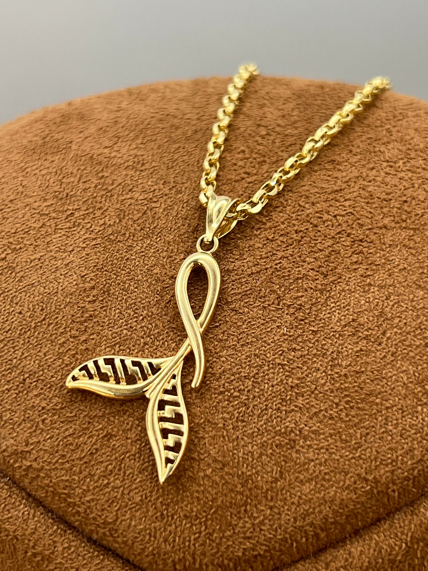 A gold necklace with a pendant combining an whale tail symbol, displayed on a brown textured jewelry bust.