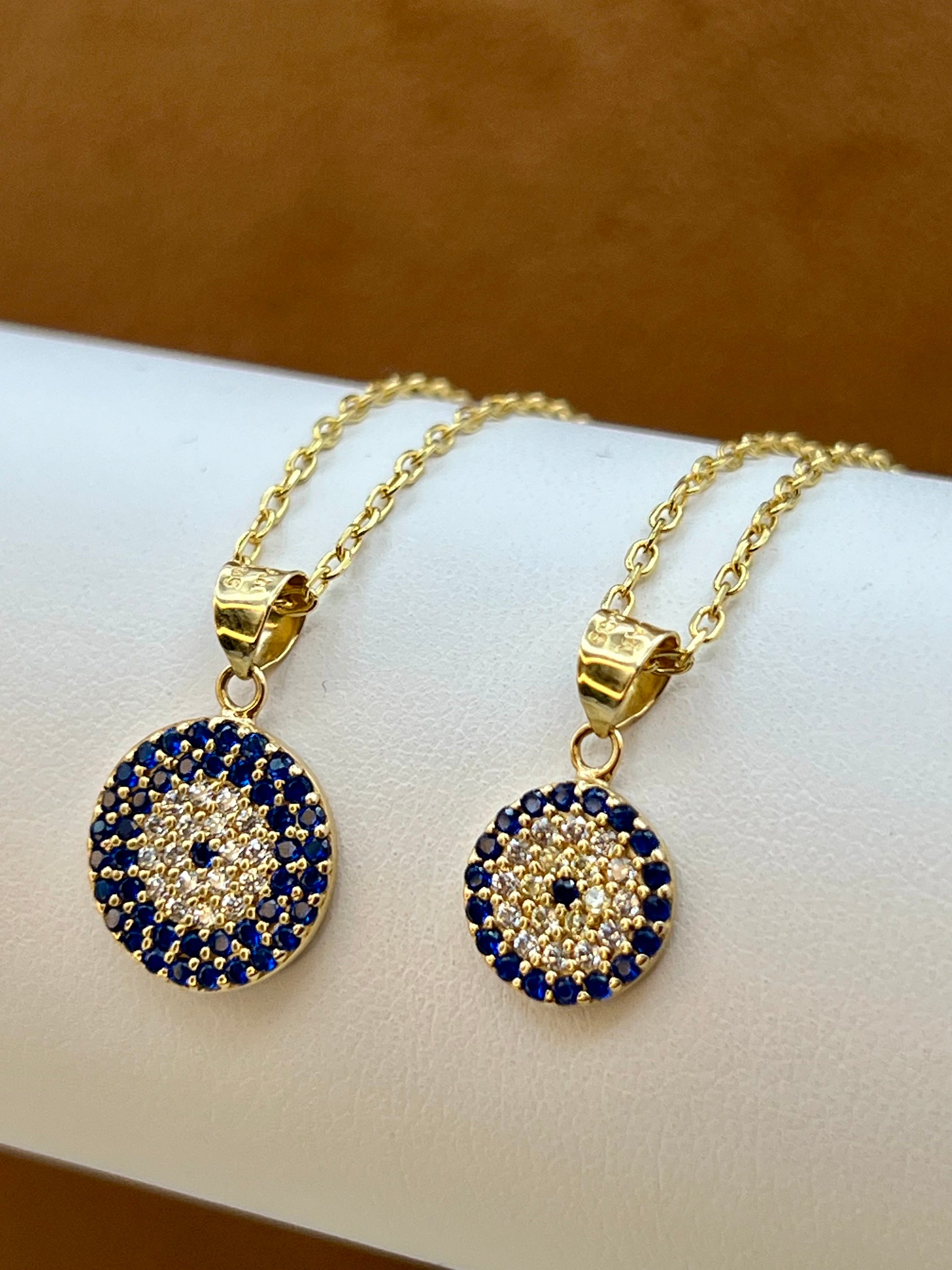 Two gold-tone necklaces with circular evil eye pendants displayed on a white jewelry stand. Each pendant features a gold border surrounded by dark blue and clear rhinestones, with a small blue center.