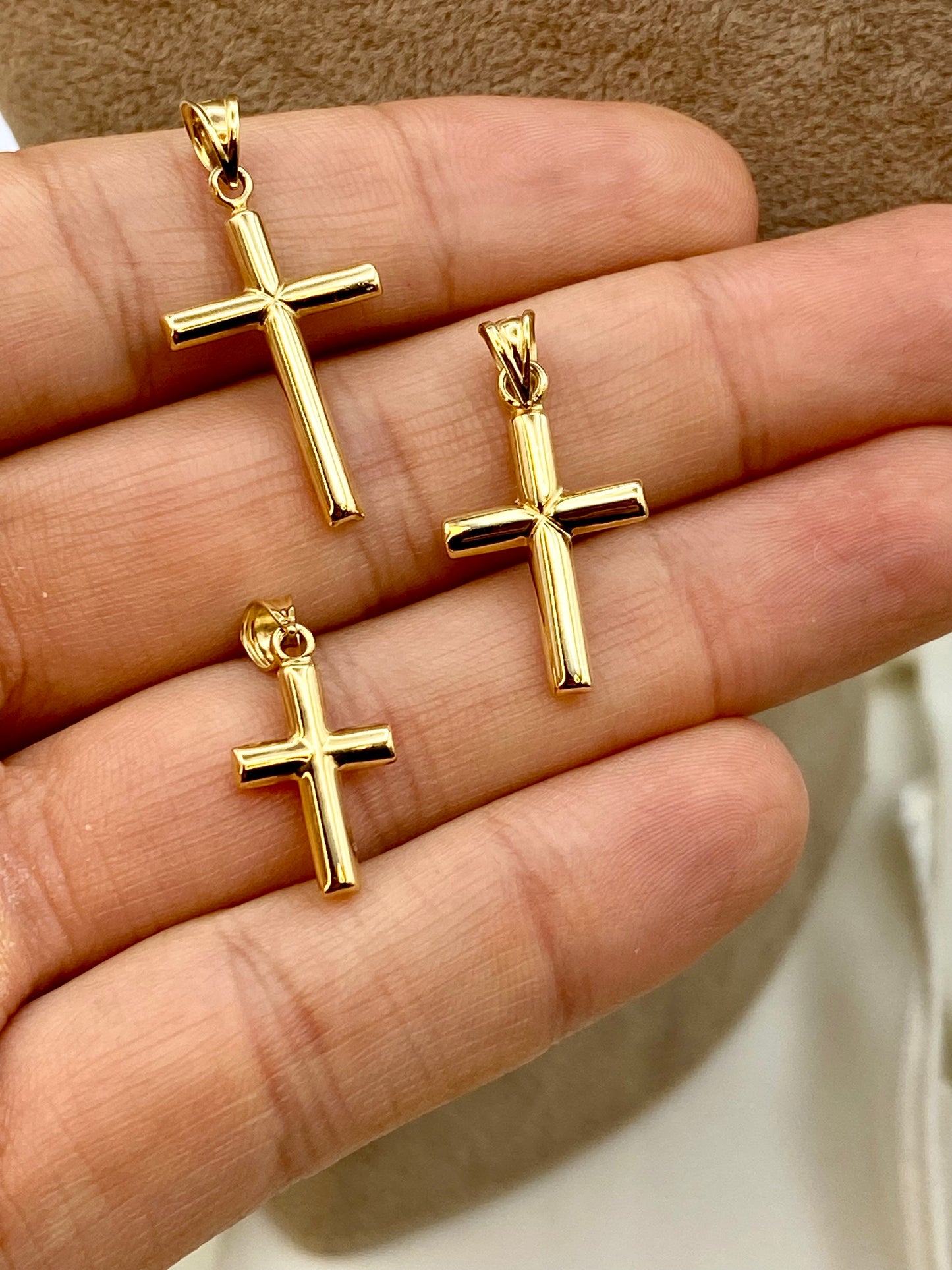 Three gold cross pendants displayed on a person's hand against a neutral background.