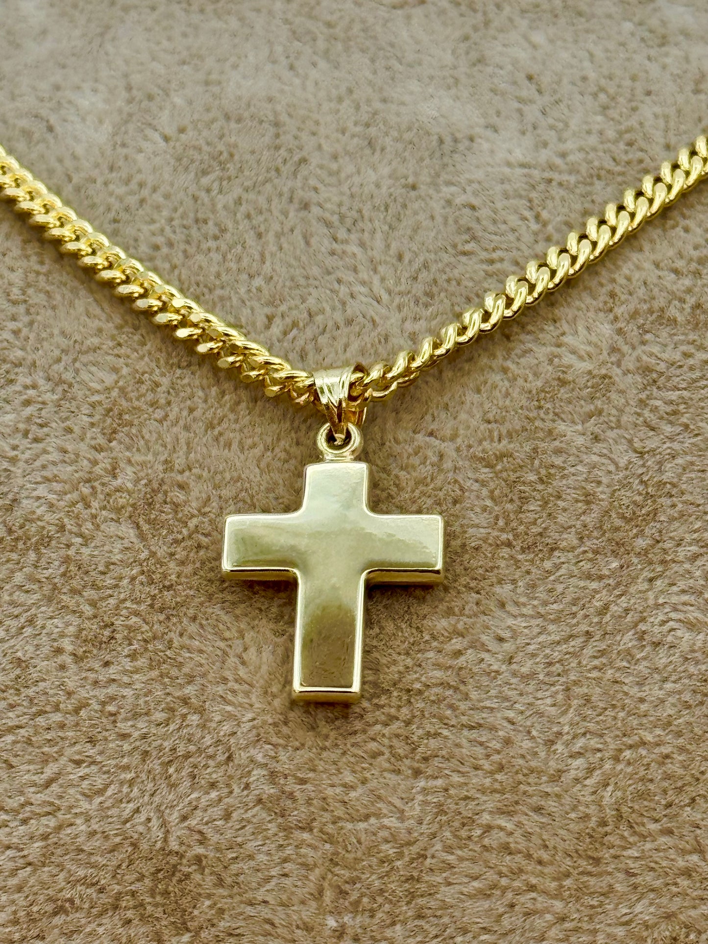 A gold cross pendant necklace on a gold chain, displayed on a textured beige fabric background.