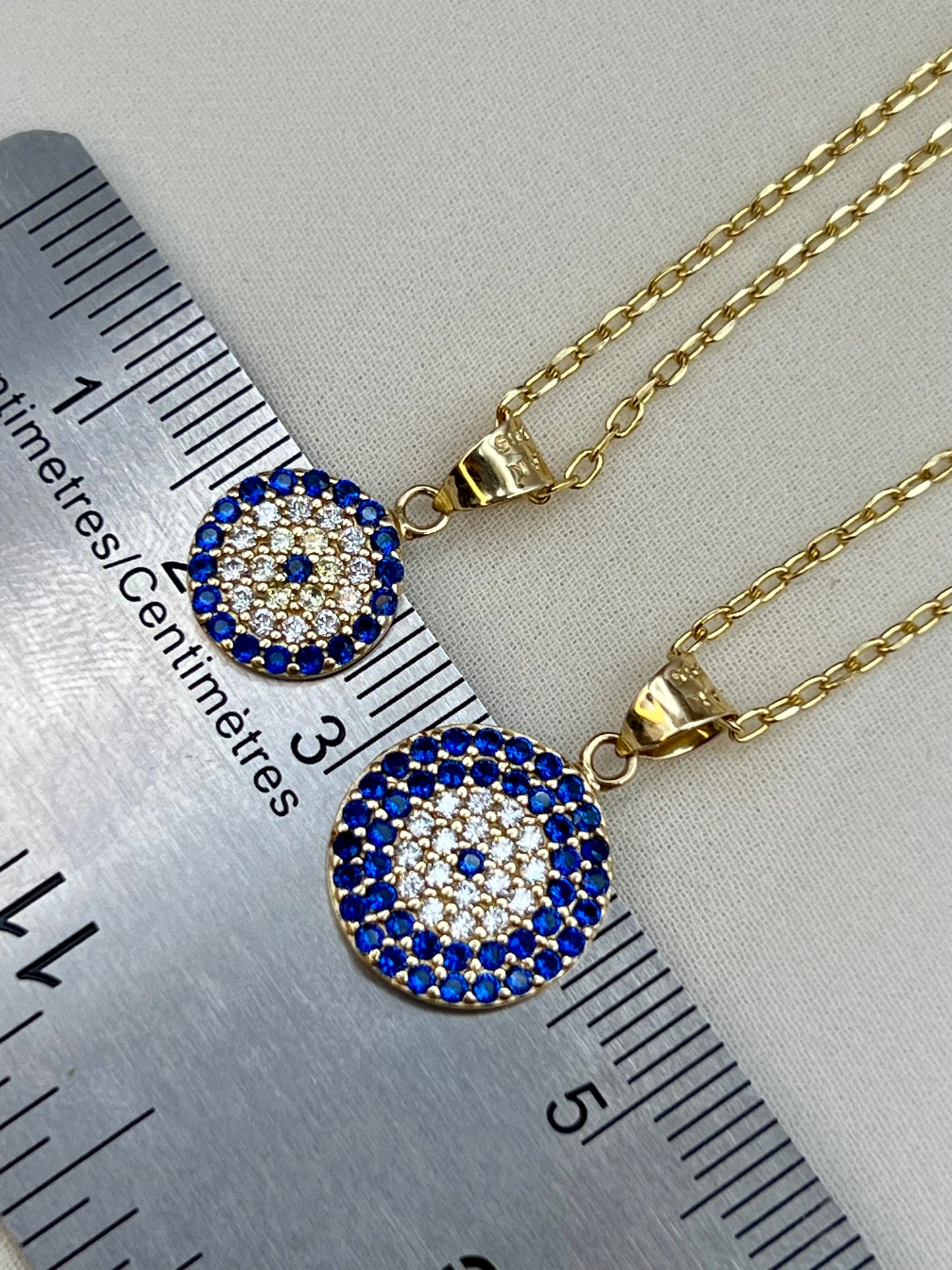 Gold-tone circular pendant necklace and matching chain with blue and white evil eye designs, shown next to a metal ruler for scale on a light fabric background.