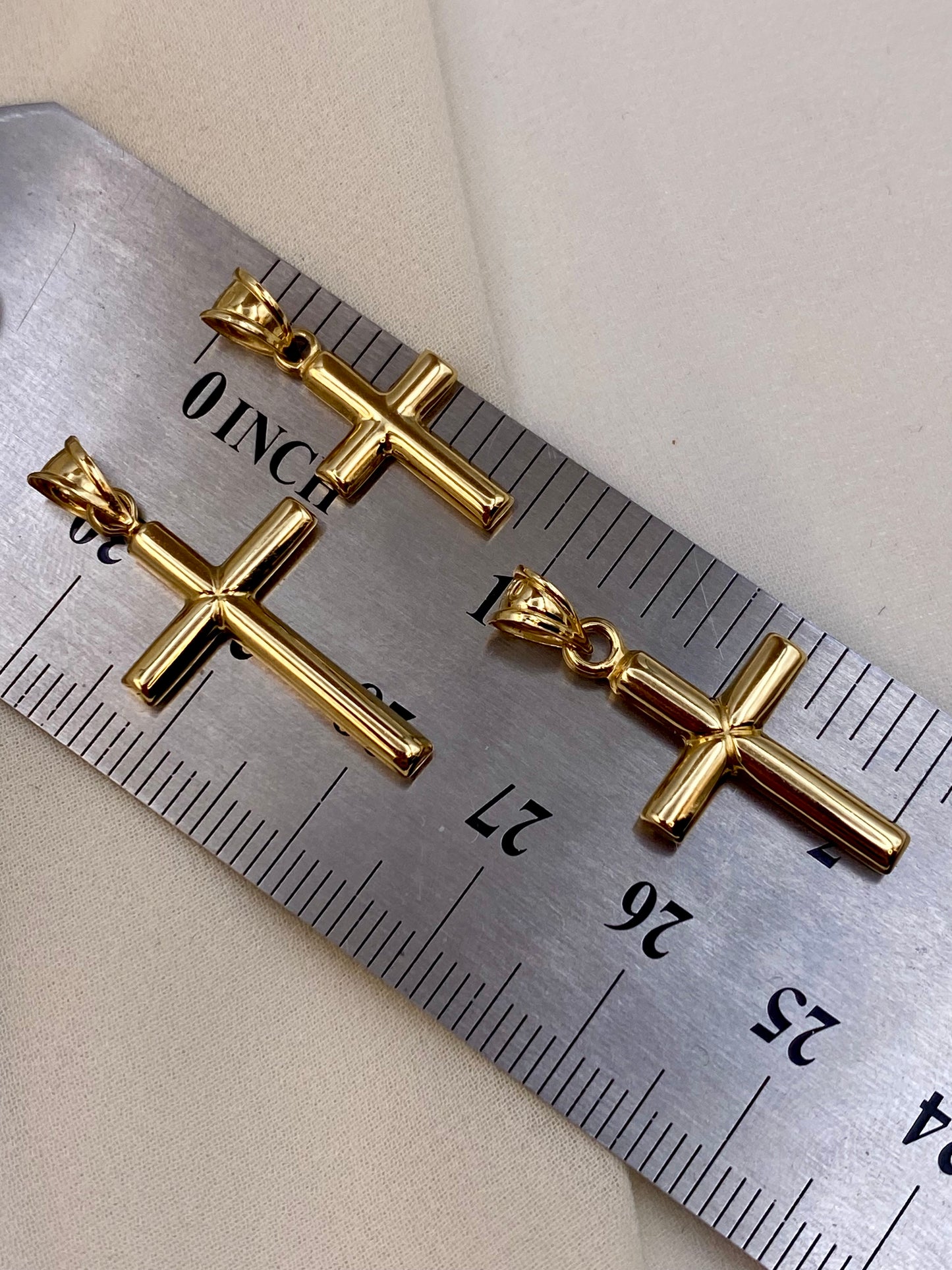 Two gold cross pendant charms displayed on a metal ruler for scale against a light beige background.