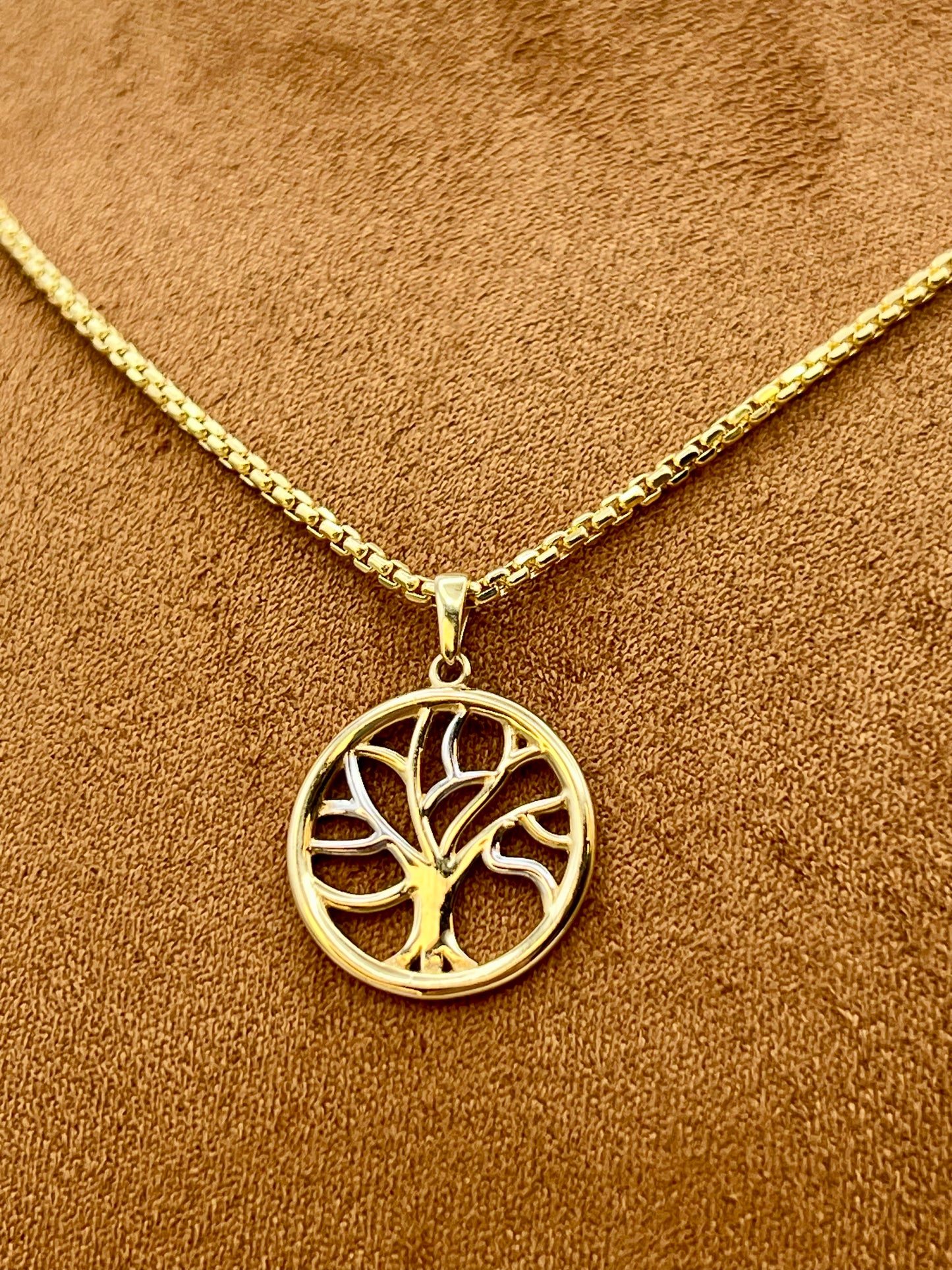 A gold tree of life pendant necklace on a matching chain, displayed on a textured brown suede-like surface.