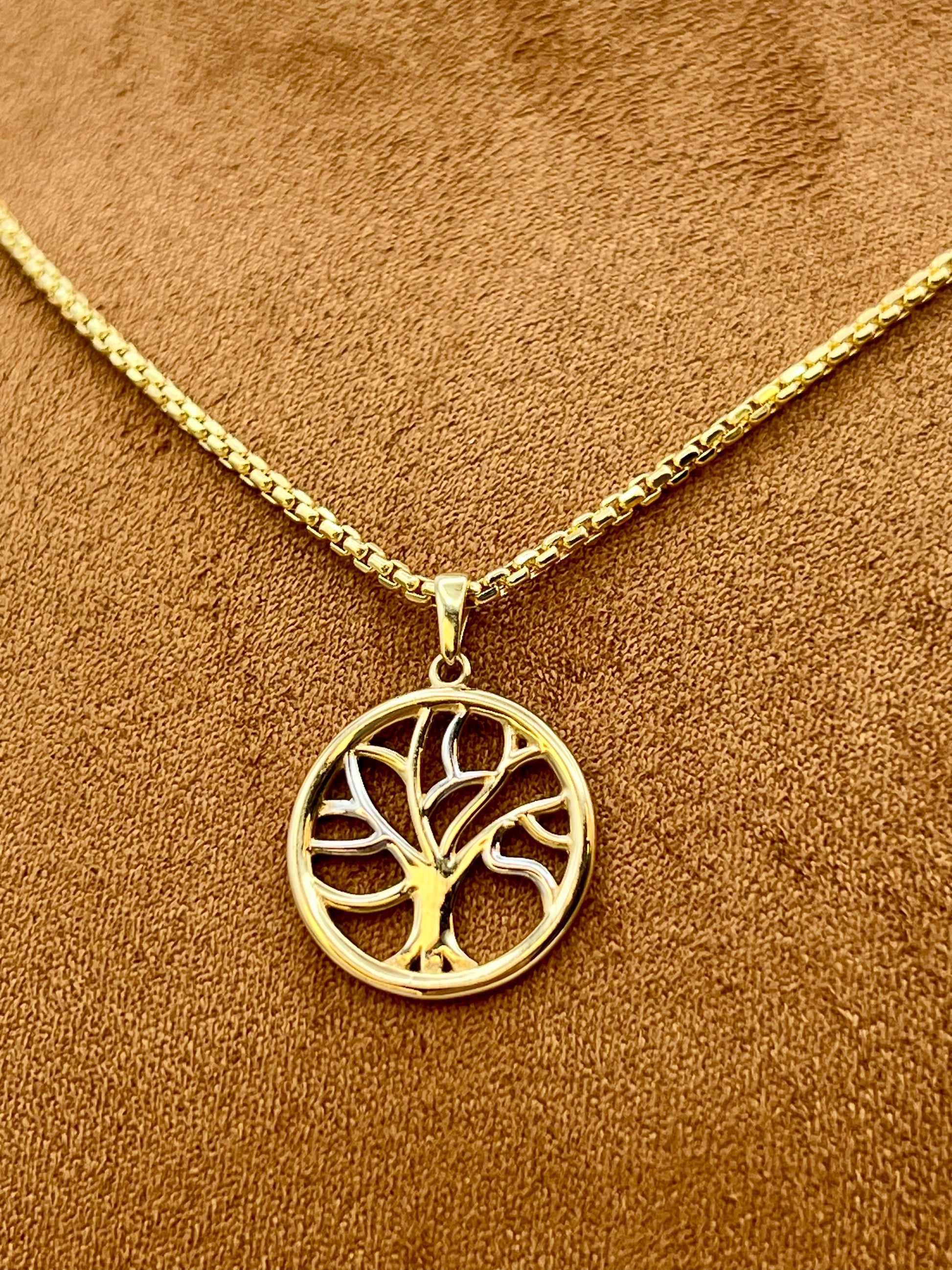 A gold tree of life pendant necklace on a matching chain, displayed on a textured brown suede-like surface.