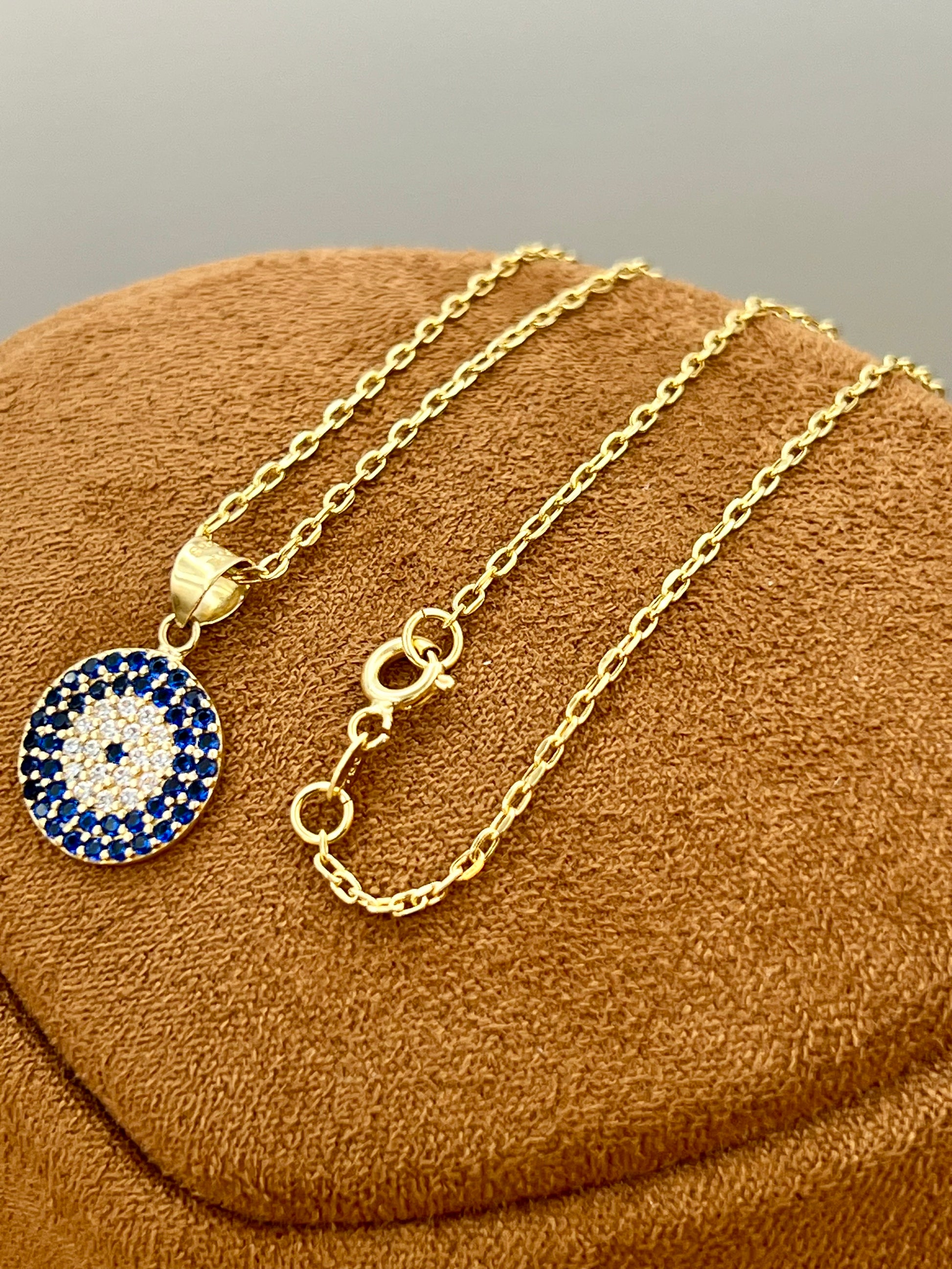 A gold chain necklace with a circular evil eye pendant, featuring a blue and white pattern, displayed on a brown textured jewelry stand.