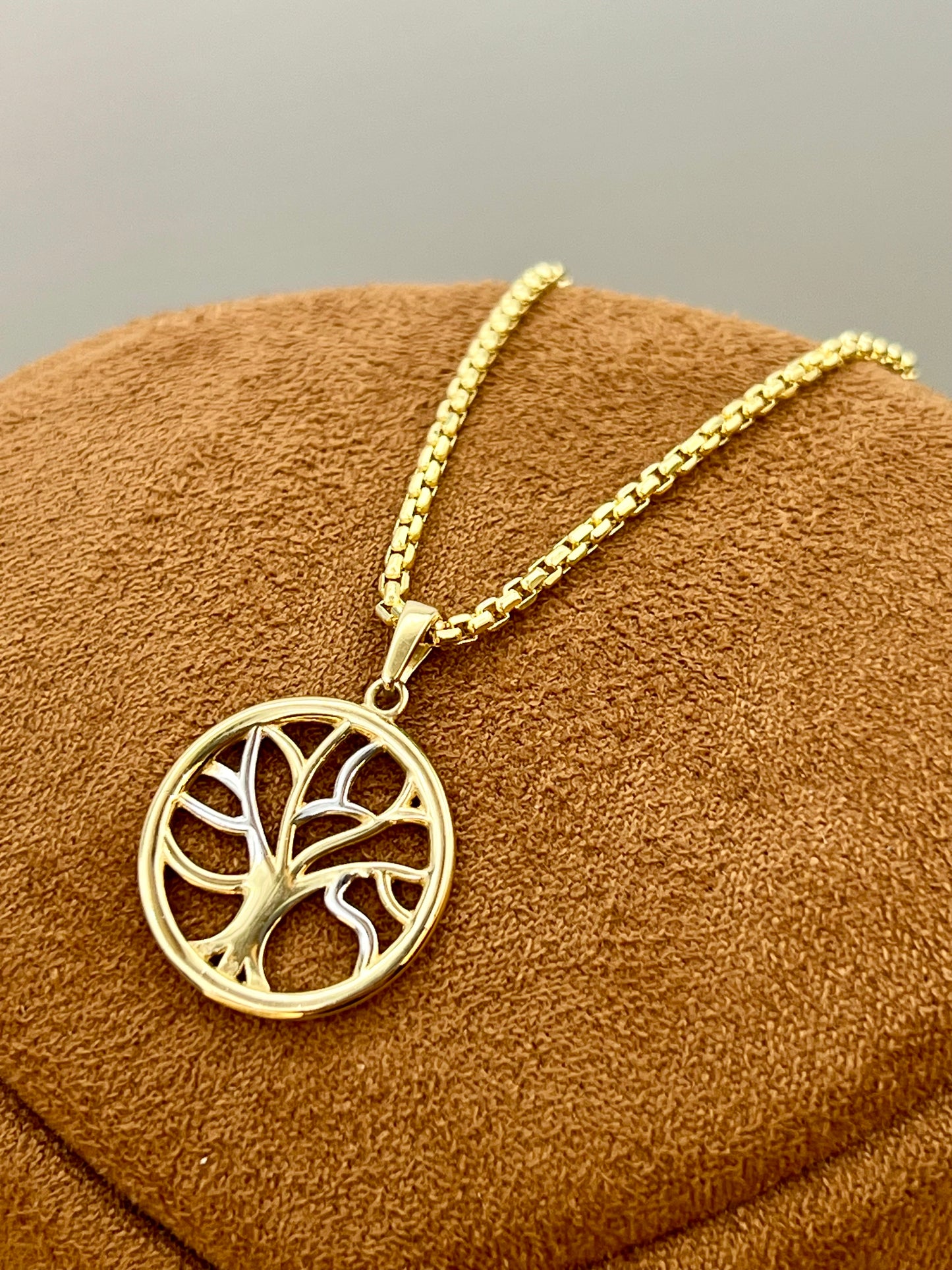 A gold tree of life pendant necklace displayed on a brown textured velvet jewelry stand against a neutral gray background.