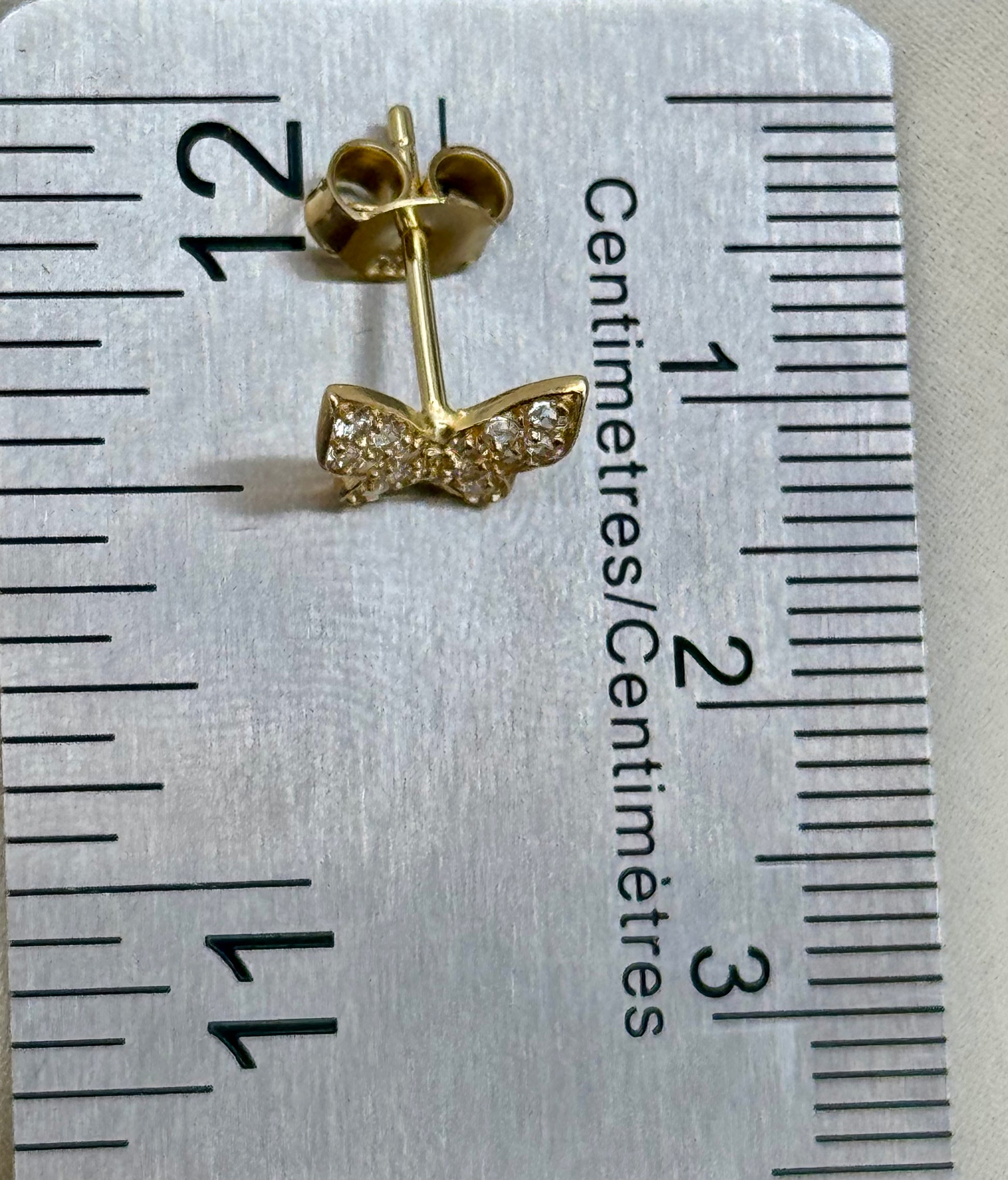 A small gold butterfly stud earring with clear pavé stones, shown on a silver ruler for scale.