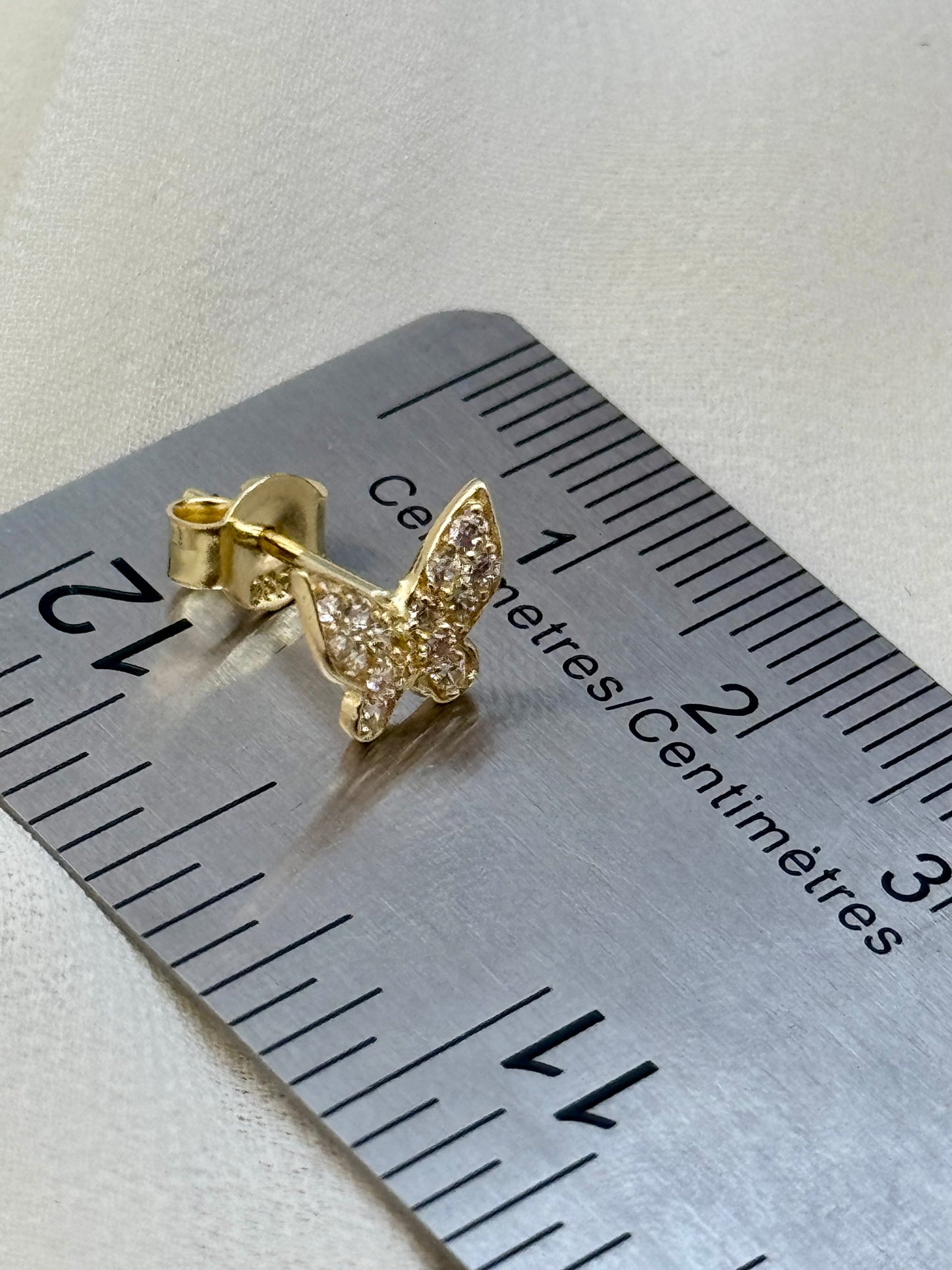 A small gold butterfly stud earring with clear pavé stones, shown on a metal ruler for scale against a light beige textured background.