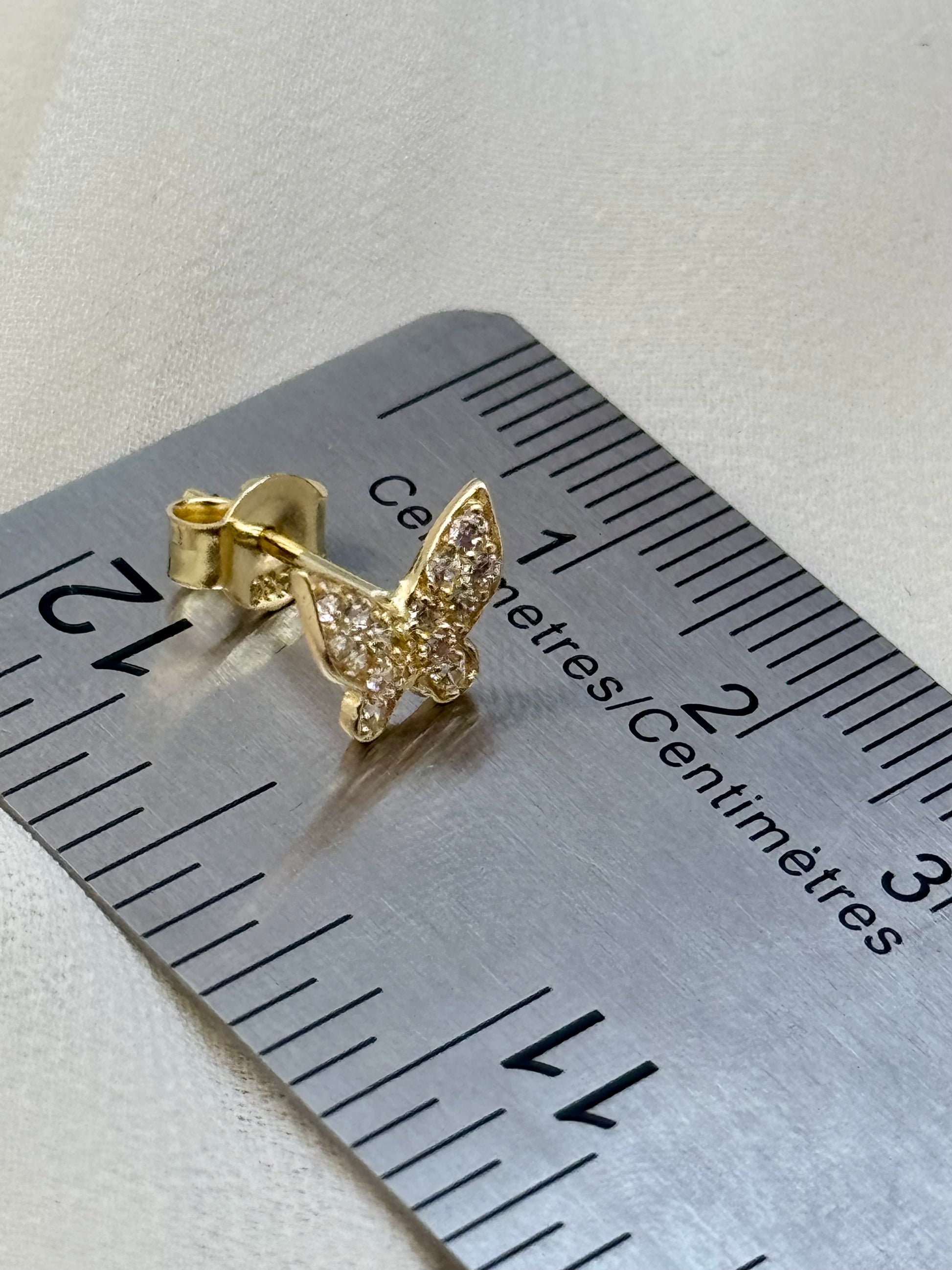 A small gold butterfly stud earring with clear pavé stones, shown on a metal ruler for scale against a light beige textured background.
