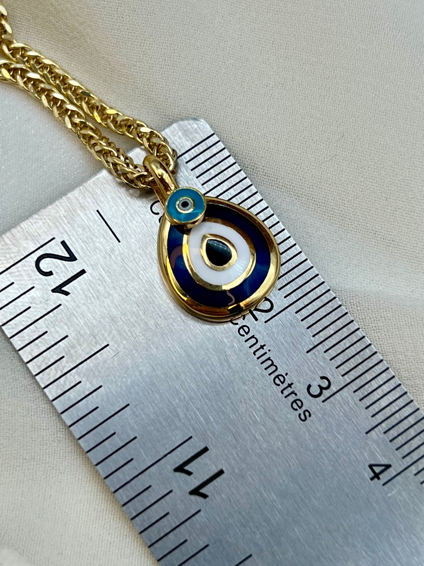 Gold-tone evil eye pendant necklace with blue and white enamel details, shown next to a metal ruler for scale on a light fabric background.