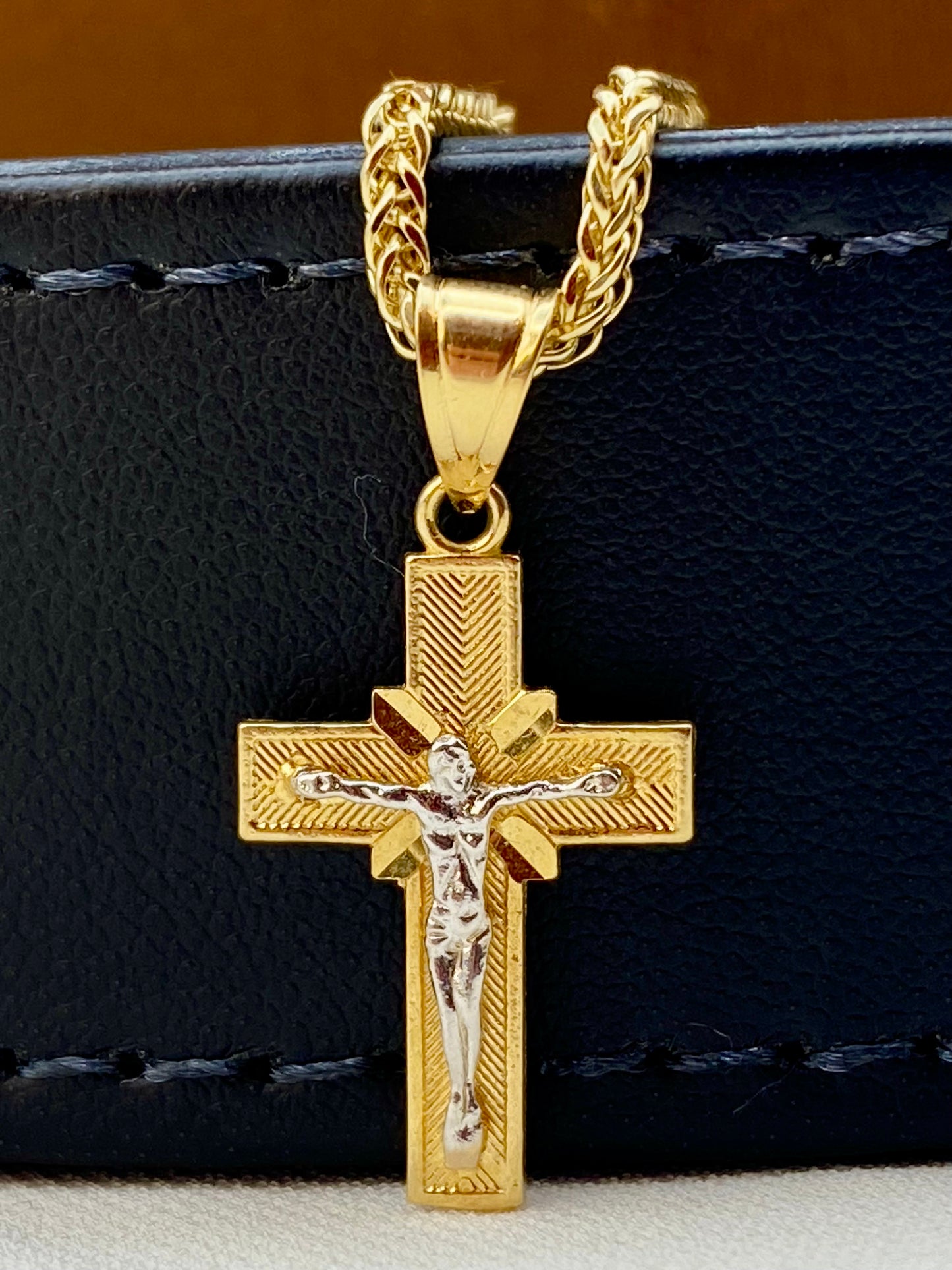 A gold cross pendant necklace with a silver-toned figure of Jesus on the cross, hanging from a gold chain against a black textured leather background.