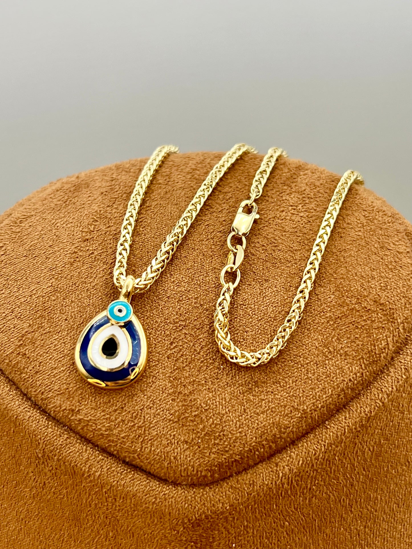 A gold chain necklace with a teardrop-shaped evil eye pendant, displayed on a brown velvet jewelry bust against a neutral gray background.