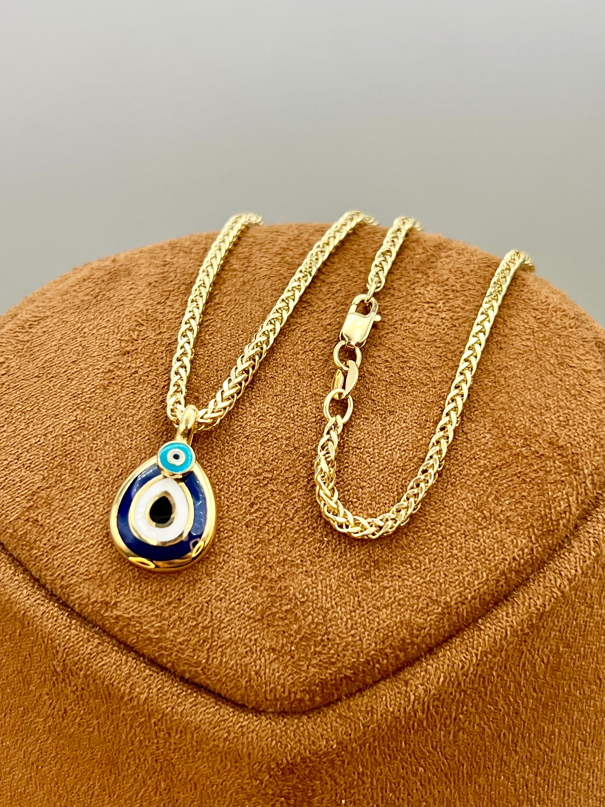 A gold chain necklace with a teardrop-shaped evil eye pendant, displayed on a brown velvet jewelry bust against a neutral gray background.