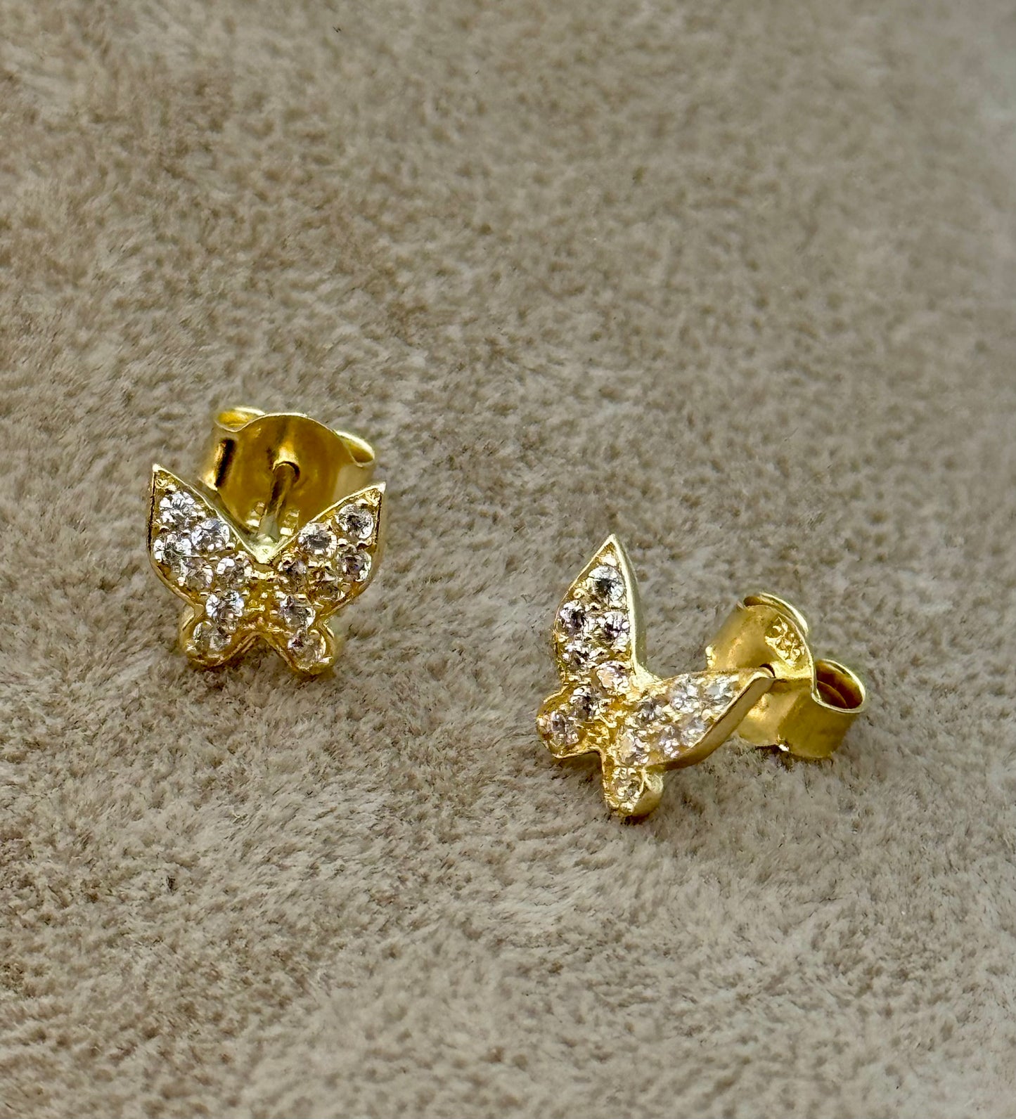 A pair of gold butterfly stud earrings adorned with small clear crystals, displayed on a textured beige surface.