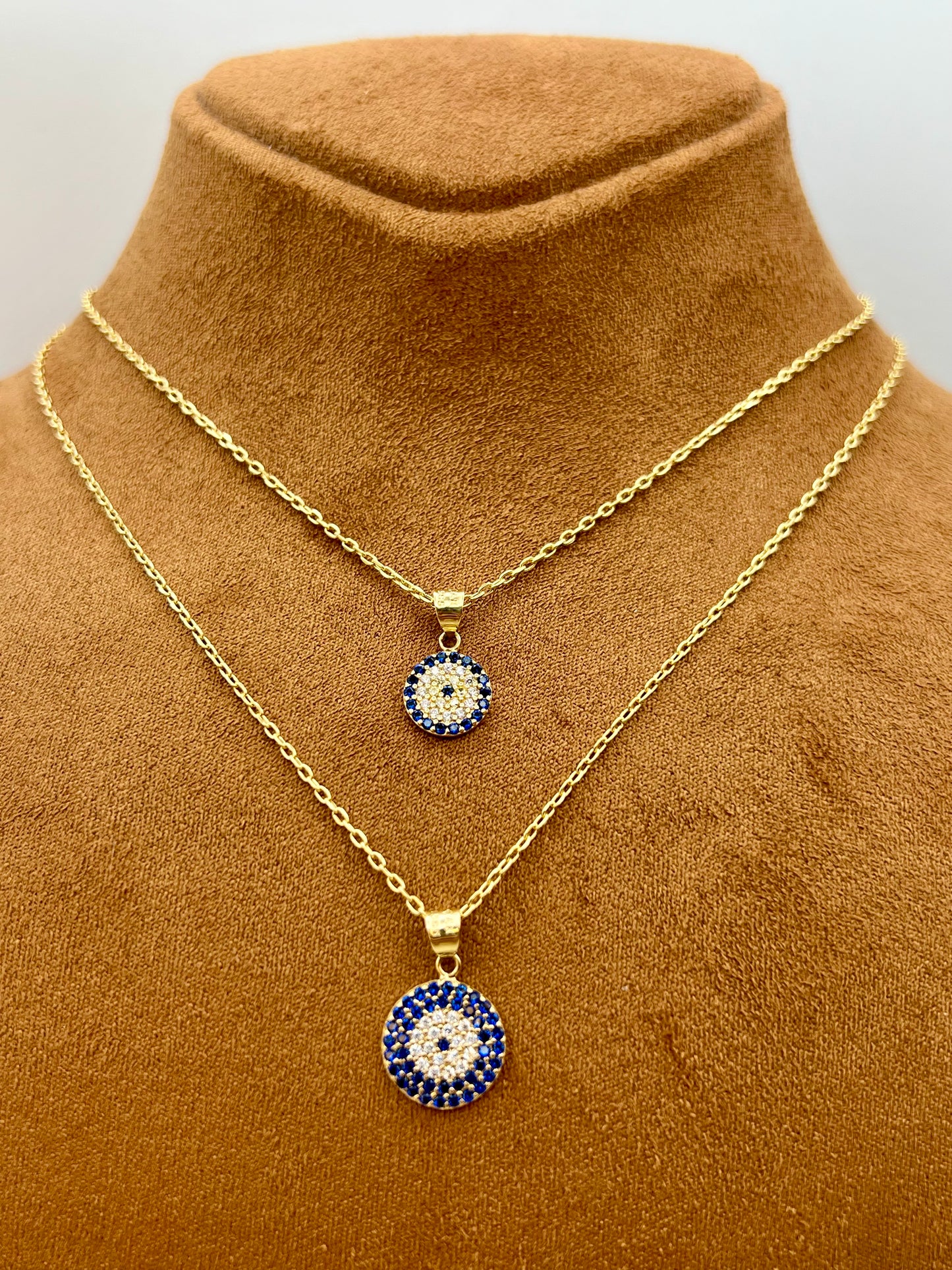 Two gold-tone necklaces displayed on a brown velvet jewelry bust. Both necklaces feature circular evil eye pendants with blue, white, and red details.