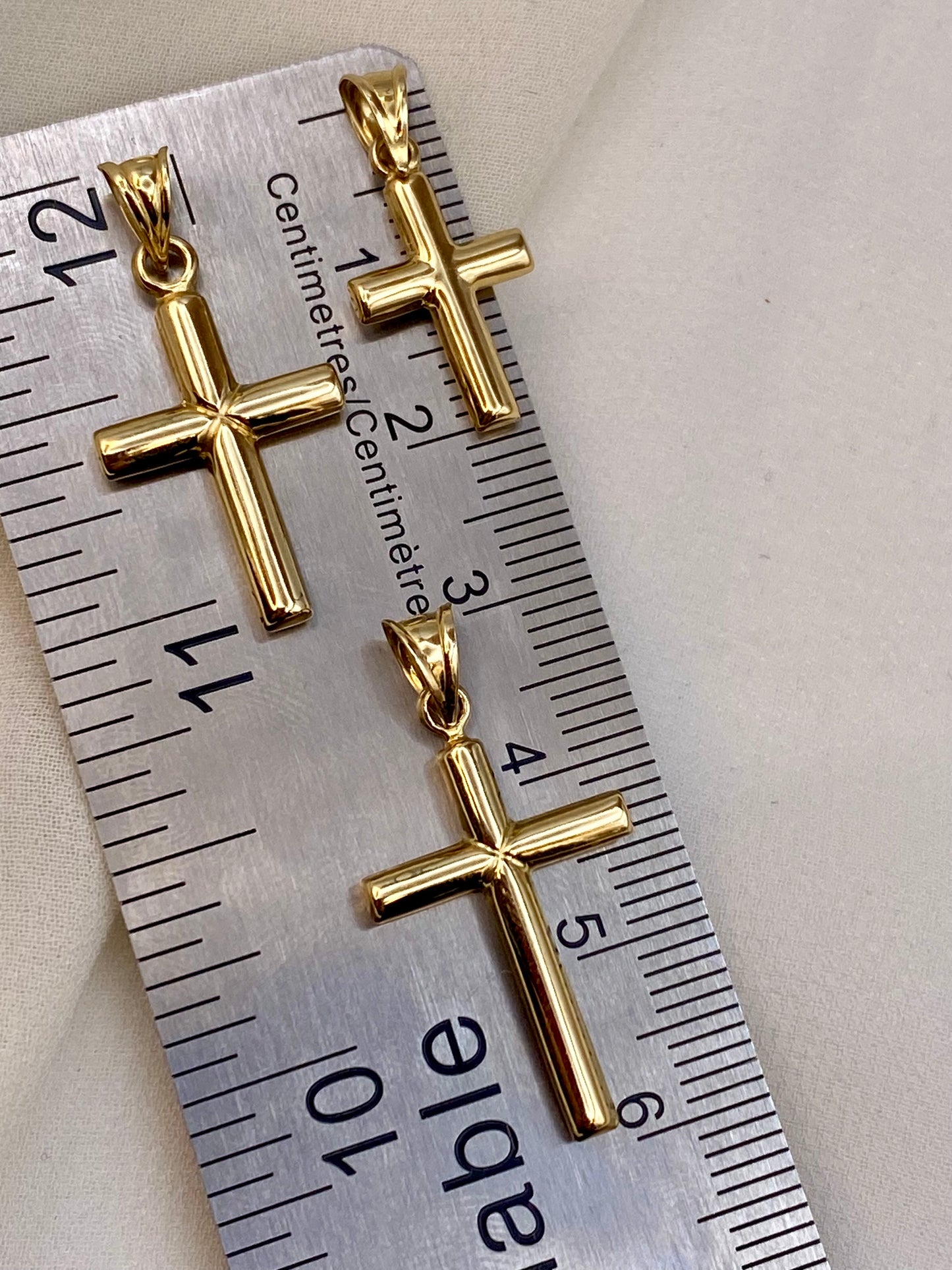 Three gold cross pendants displayed on a silver metal ruler for scale, set against a light beige fabric background.