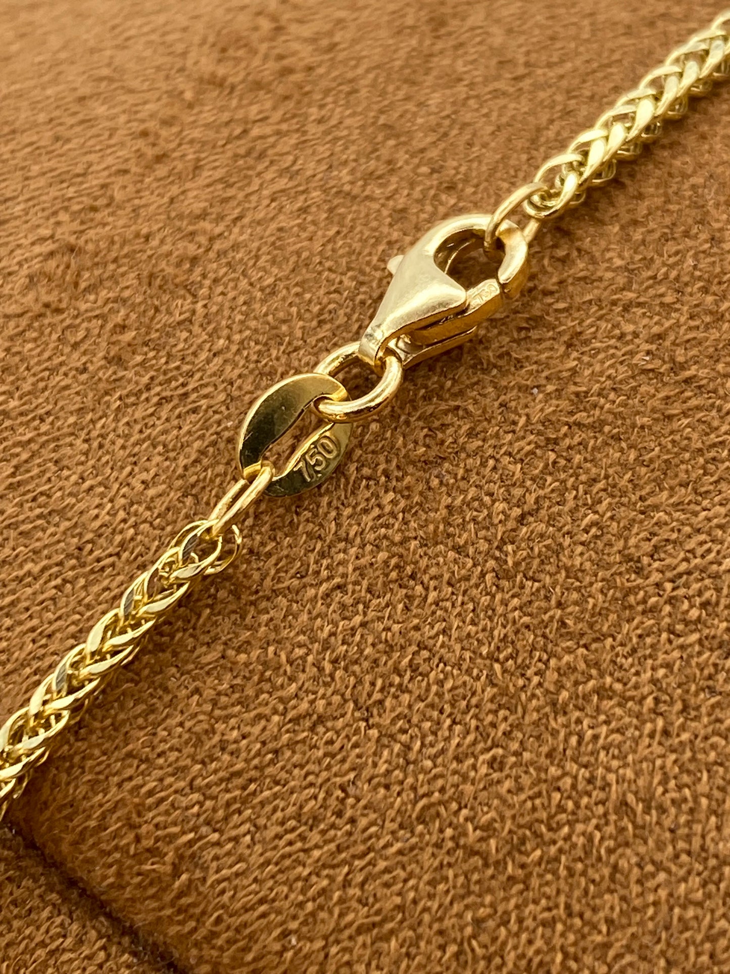 Close-up of a gold chain bracelet with a lobster clasp and a round 14k gold hallmark tag, resting on a textured brown suede surface.